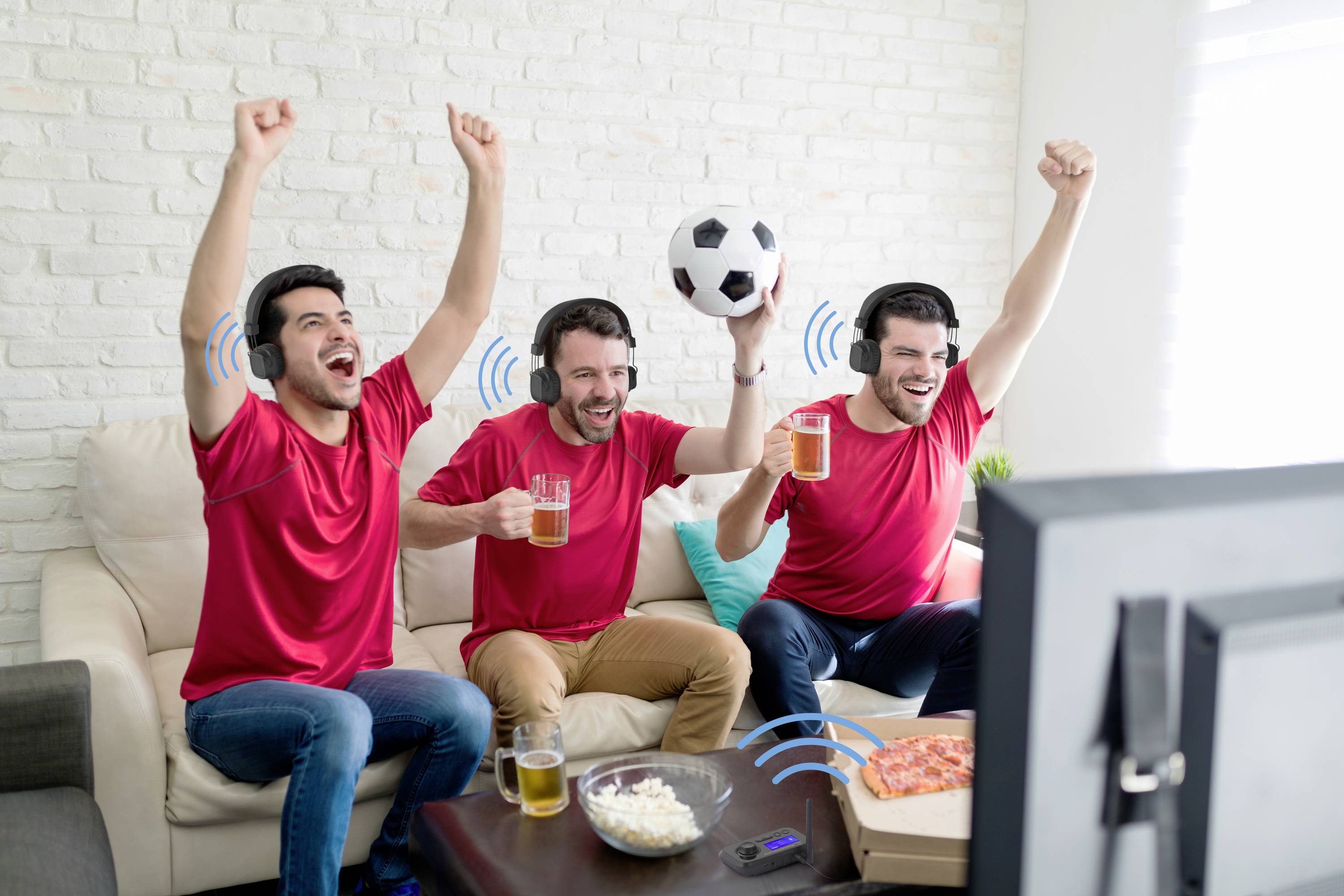 Drie mannen in rode shirts juichen voor een tv, houden drankjes en een voetbal vast terwijl ze een wedstrijd bekijken.