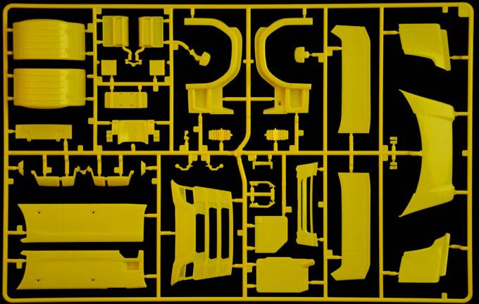 Gele kunststof gietframe met vormen van auto-onderdelen op een zwarte achtergrond, toont verschillende componenten voor de assemblage van een modelauto.