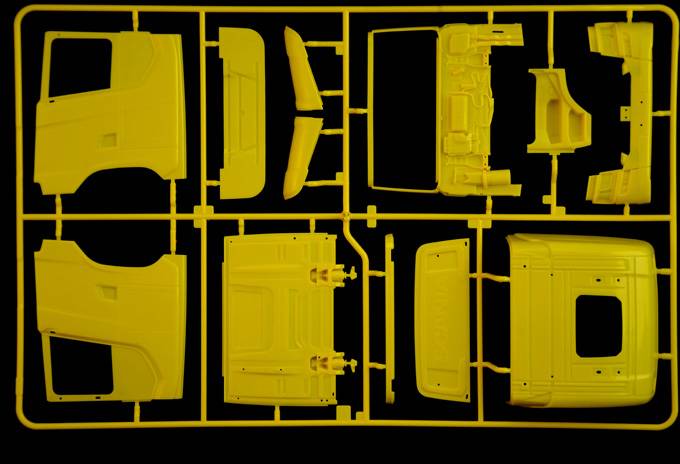Een gele kunststof bouwset met verschillende onderdelen van een modelvoertuig, gerangschikt op een zwarte achtergrond.