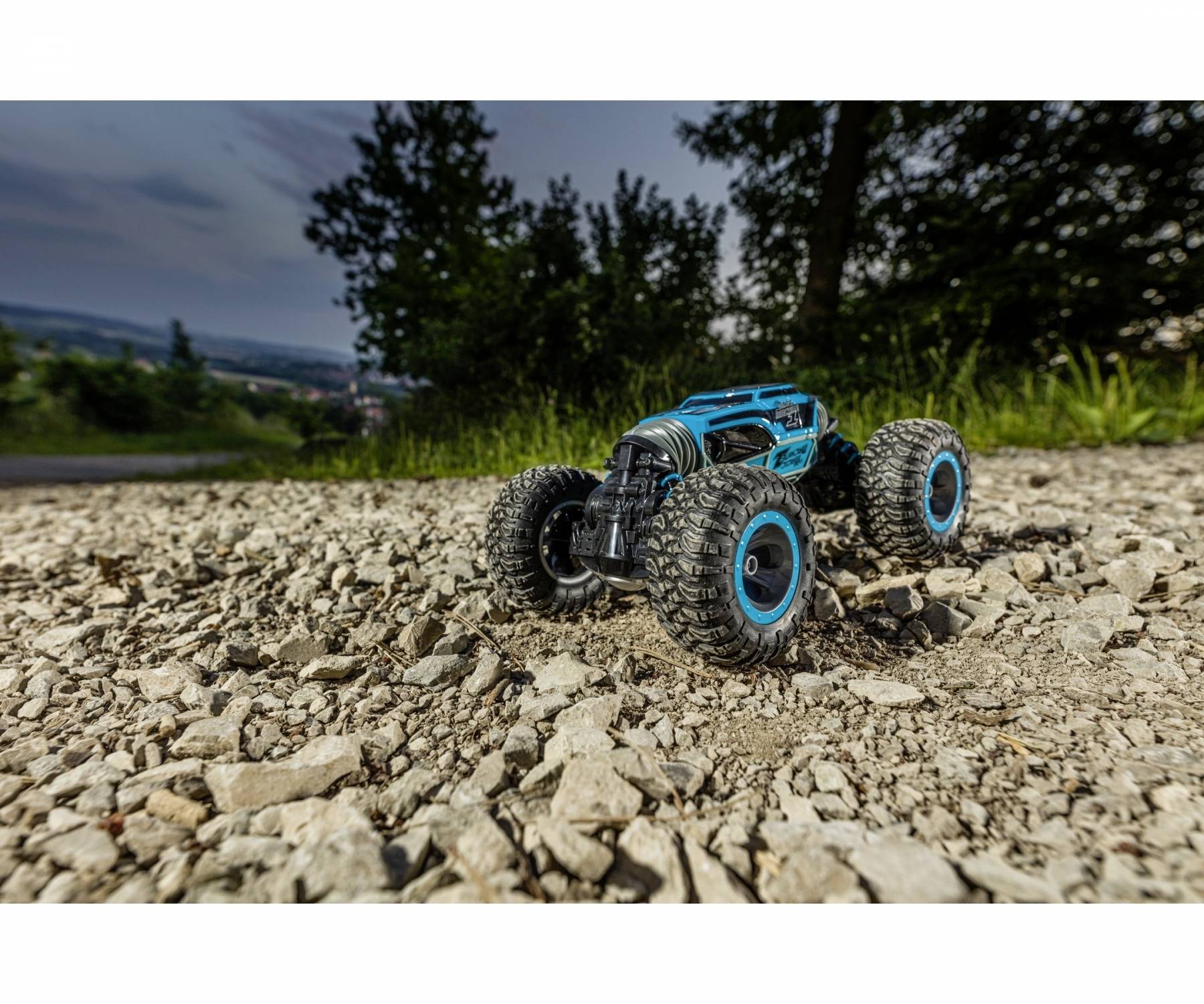 Een blauwe op afstand bestuurde terreinwagen met grote banden rijdt over een grindpad. Op de achtergrond zijn bomen en lucht zichtbaar.