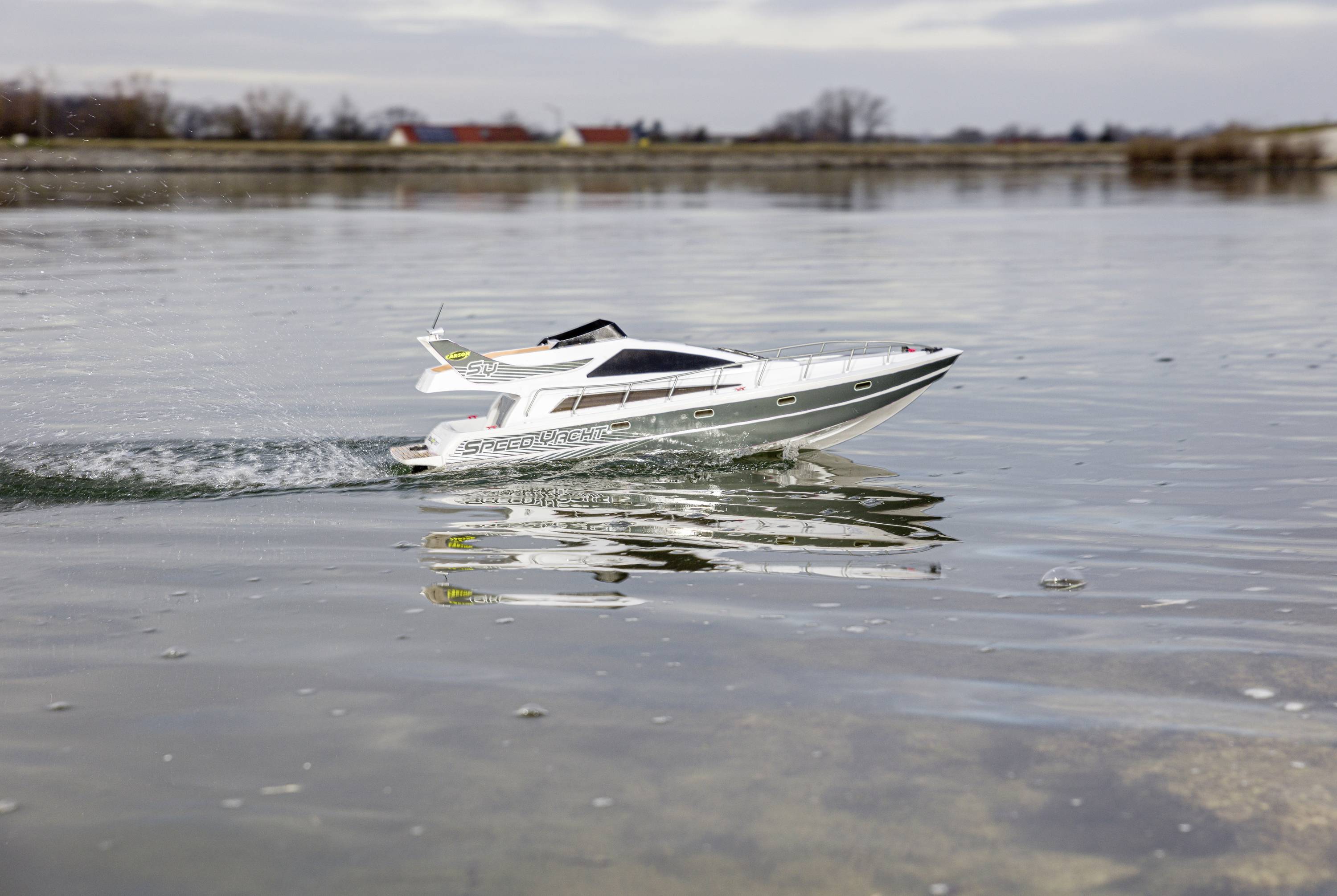 Een kleine afstandsbediende modelboot vaart op een rustig wateroppervlak. Op de achtergrond zijn enkele bomen en huizen wazig zichtbaar.