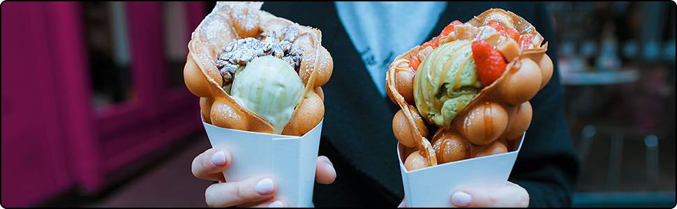 Twee handen houden wafelkegels vast met ronde bubbels. Elke wafel is gevuld met roomijs en bedekt met fruit. Achtergrond is onscherp.