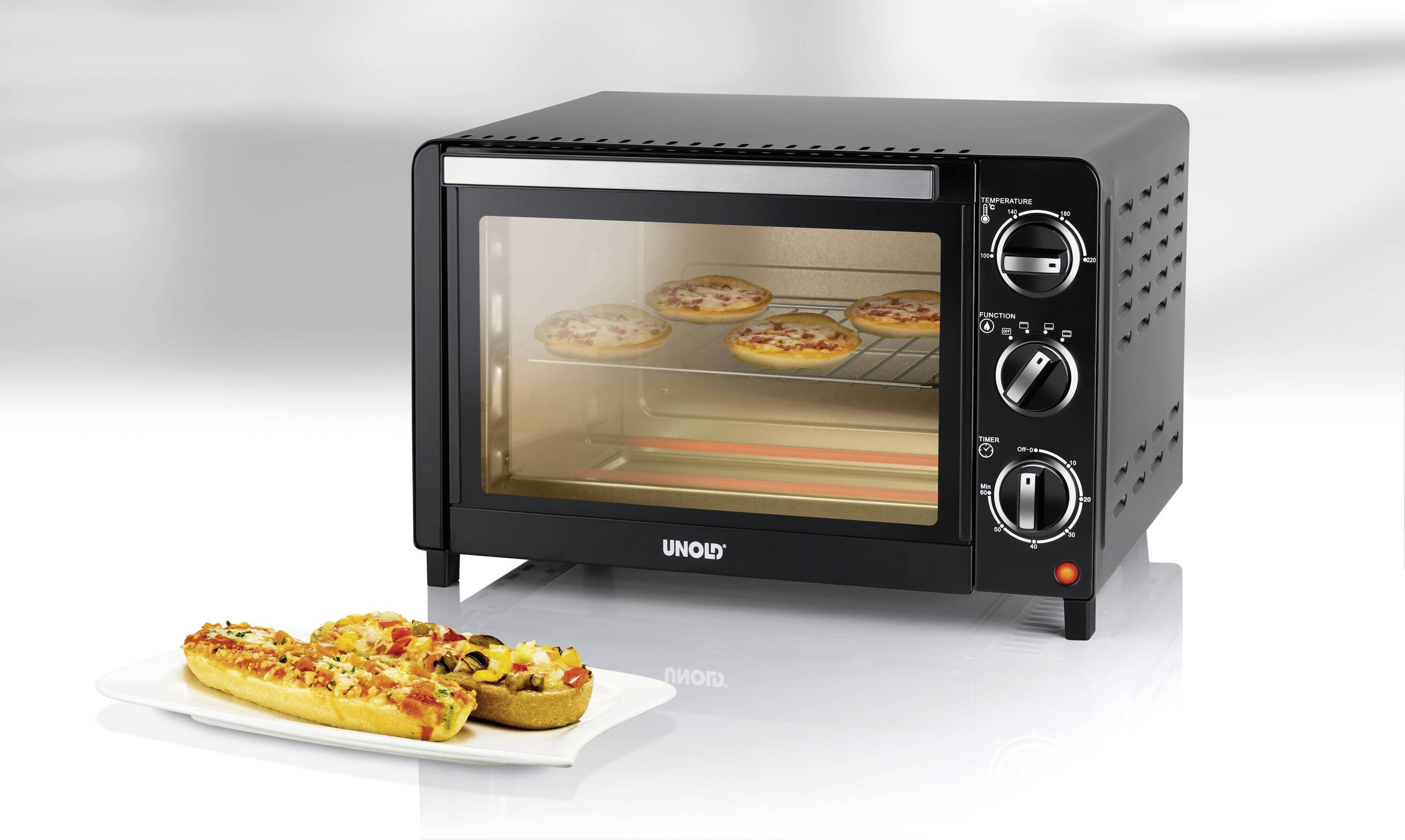 Zwarte mini-oven met glazen schuifdeur, met kleine pizza's op een rooster. Ernaast op een bord gegratineerde broden.