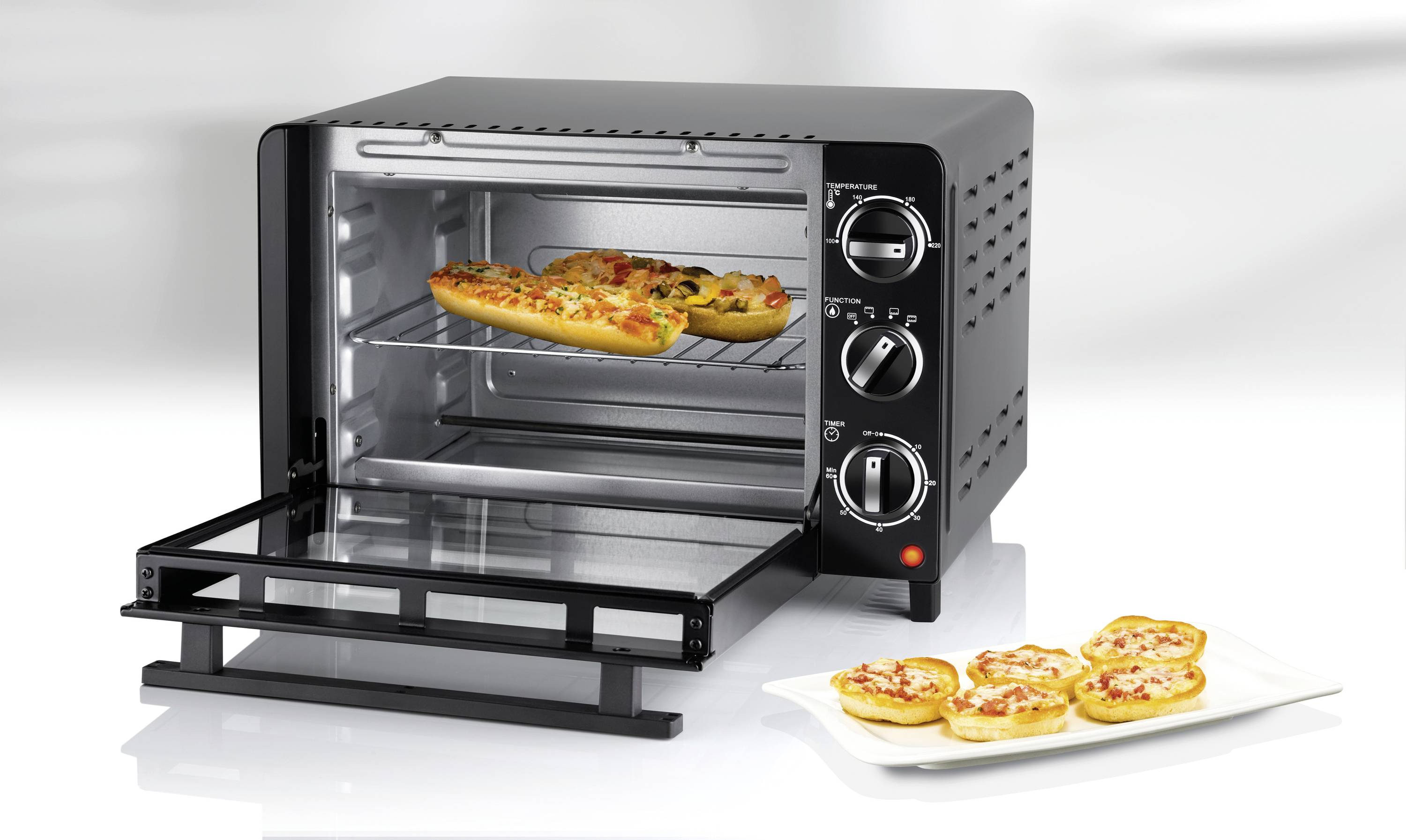 Een zwarte mini-oven met geopende deur. In de oven bevinden zich twee kleine pizza's op een bakplaat, ernaast een bord met mini-pizza's.