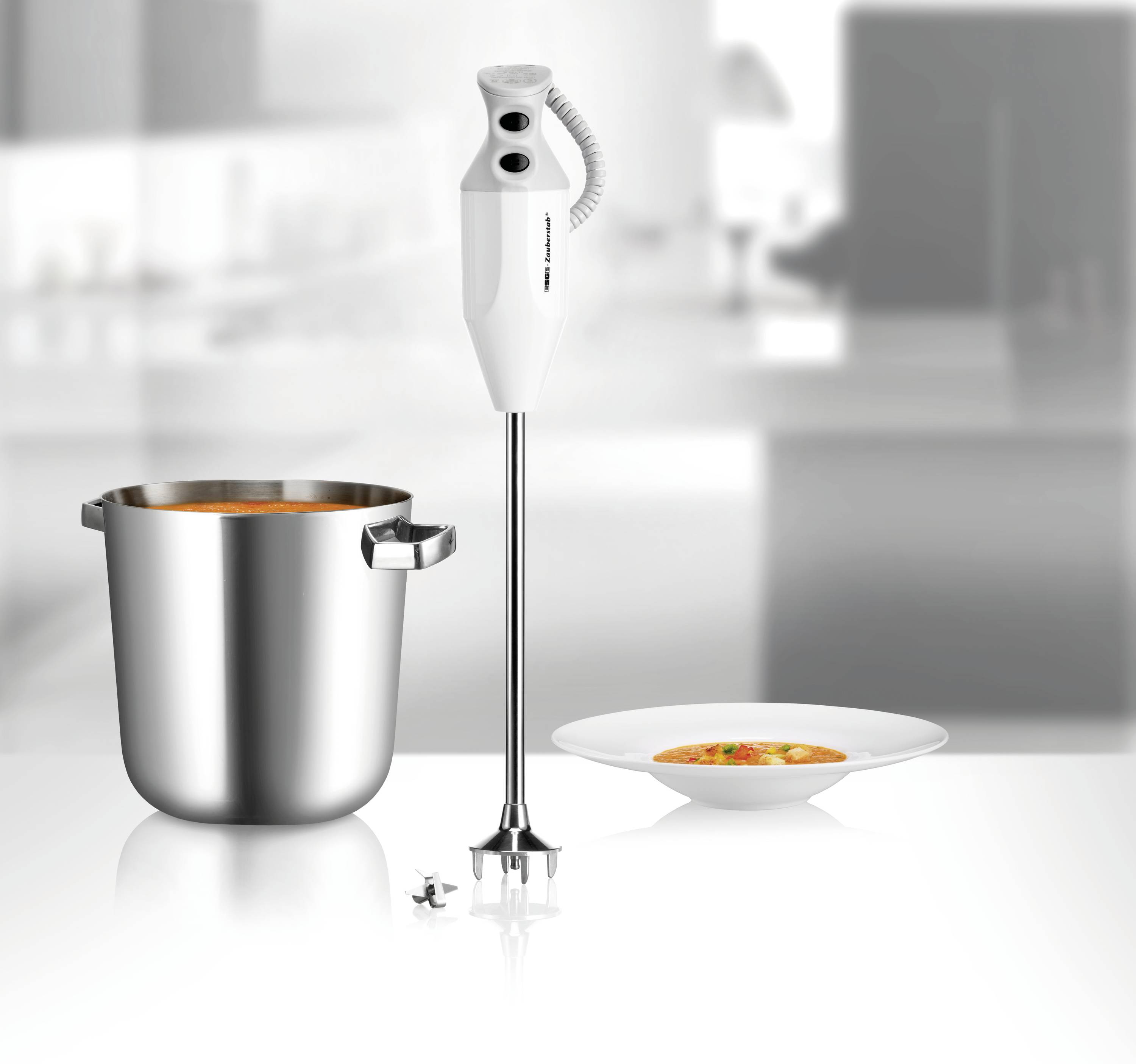 Staafmixer naast een pan met soep en een gevulde bord op een tafel in een moderne keuken.