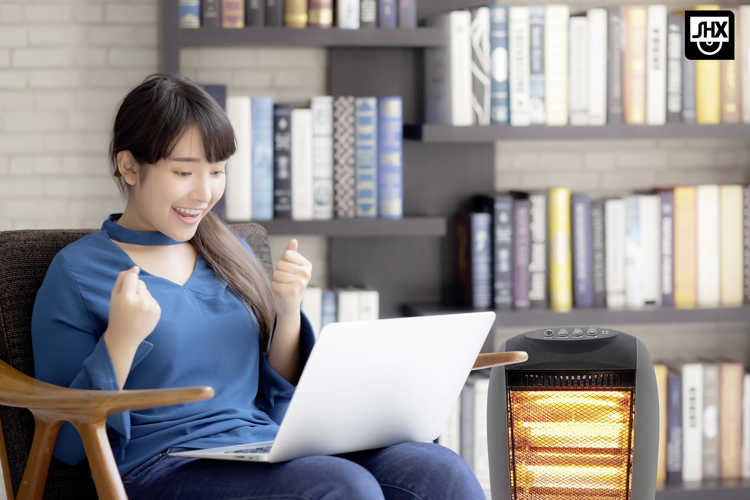 Een vrouw zit enthousiast met een laptop op een fauteuil. Op de achtergrond staat een boekenplank en rechts staat een verwarmingstoestel.