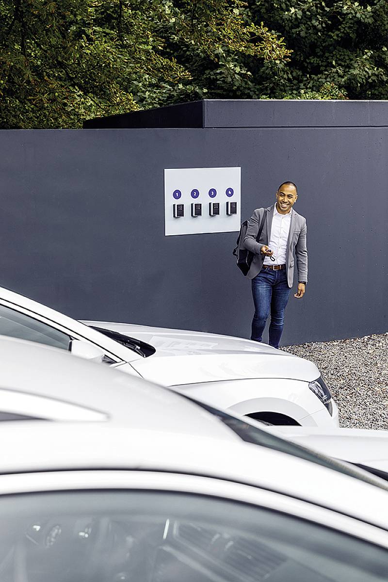 Een persoon in formele kleding glimlacht terwijl hij of zij langs een rij laadstations voor elektrische auto's loopt. Op de voorgrond zijn auto's zichtbaar.