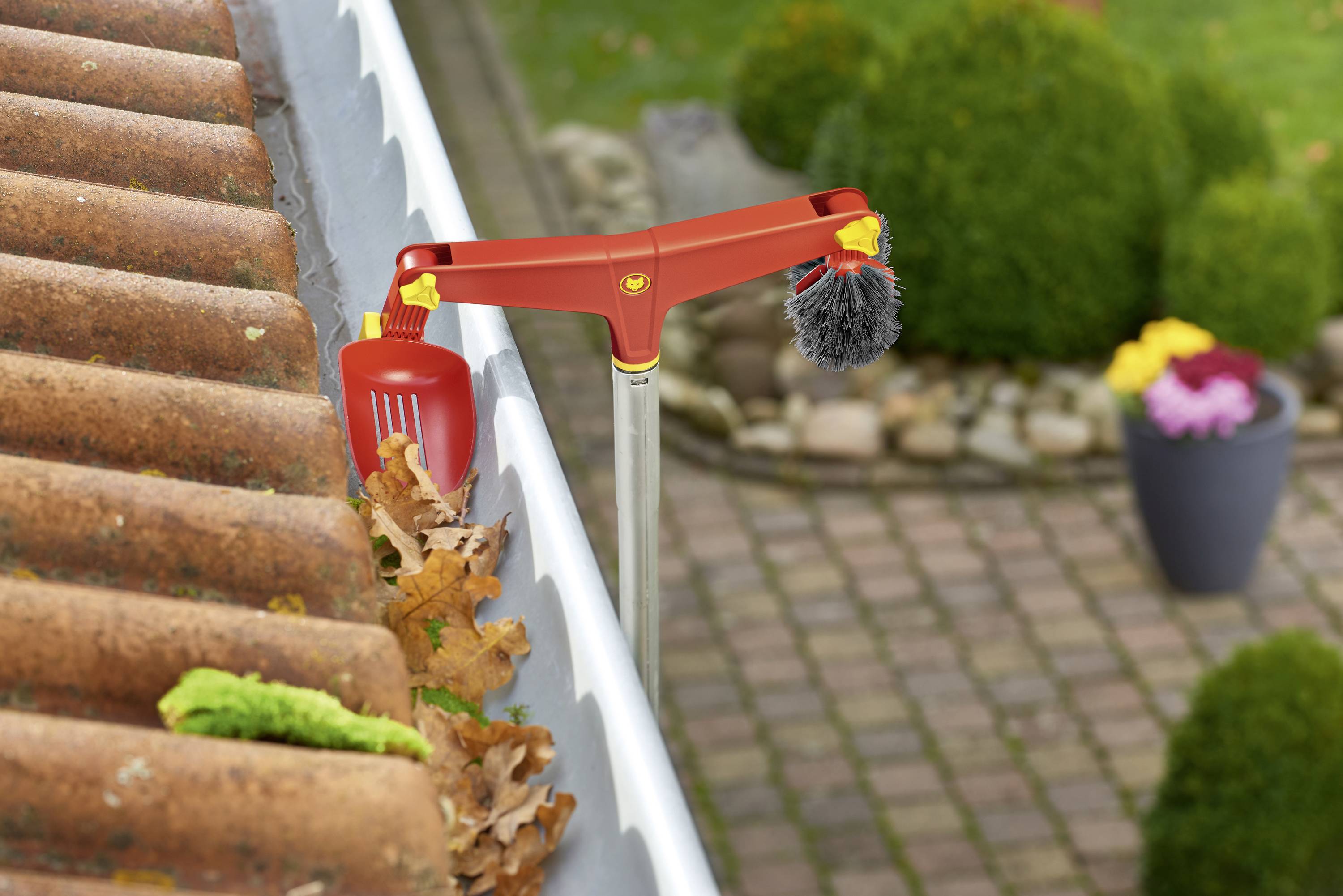Een rood reinigingsapparaat verwijdert herfstbladeren uit een dakgoot nabij baksteenrode dakpannen; planten op de achtergrond.