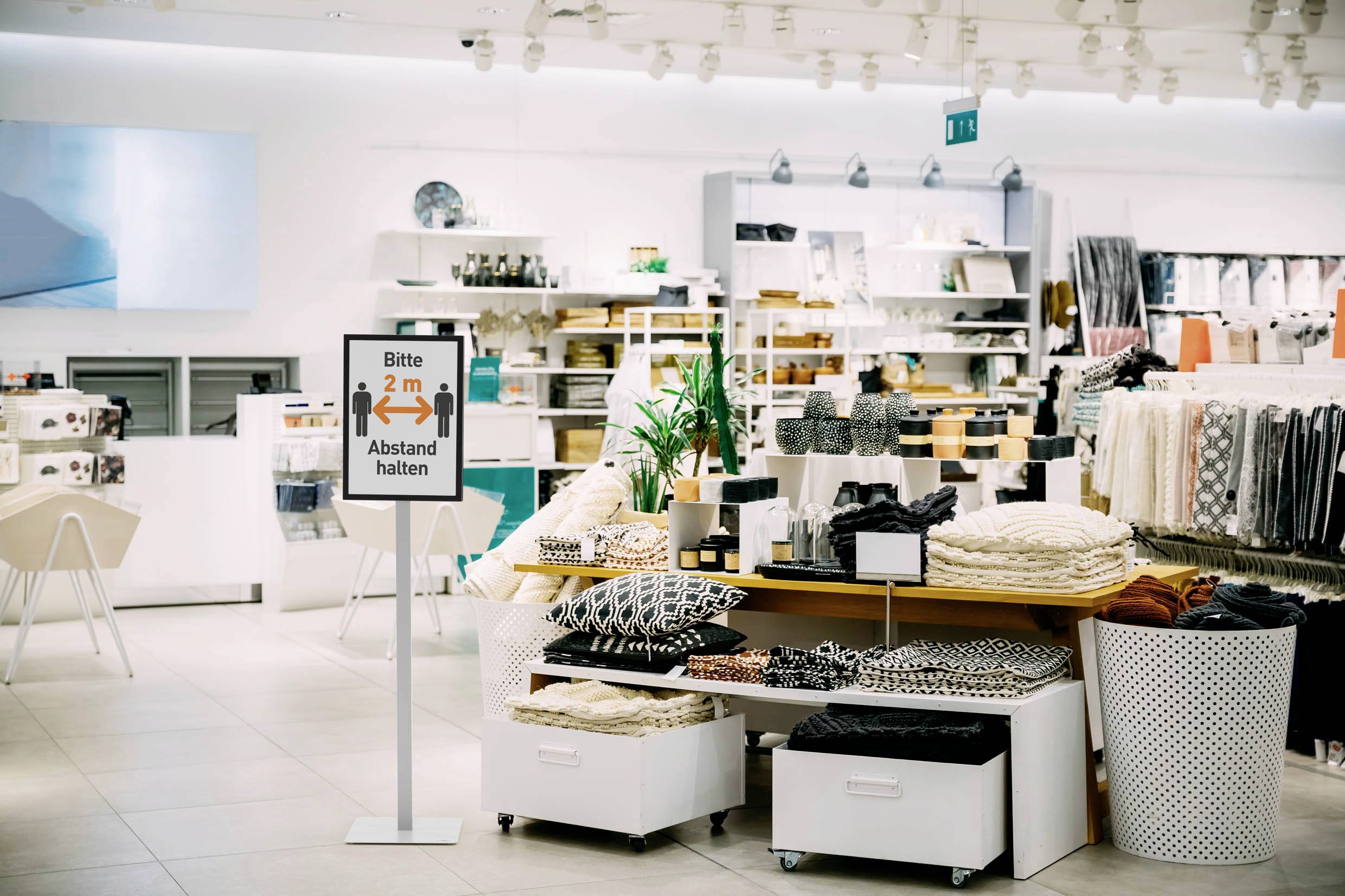 Een huishoudwinkel met een bordje: 'Gelieve 2 meter afstand te houden'. Schappen vol met kussens, textiel en decoratie.
