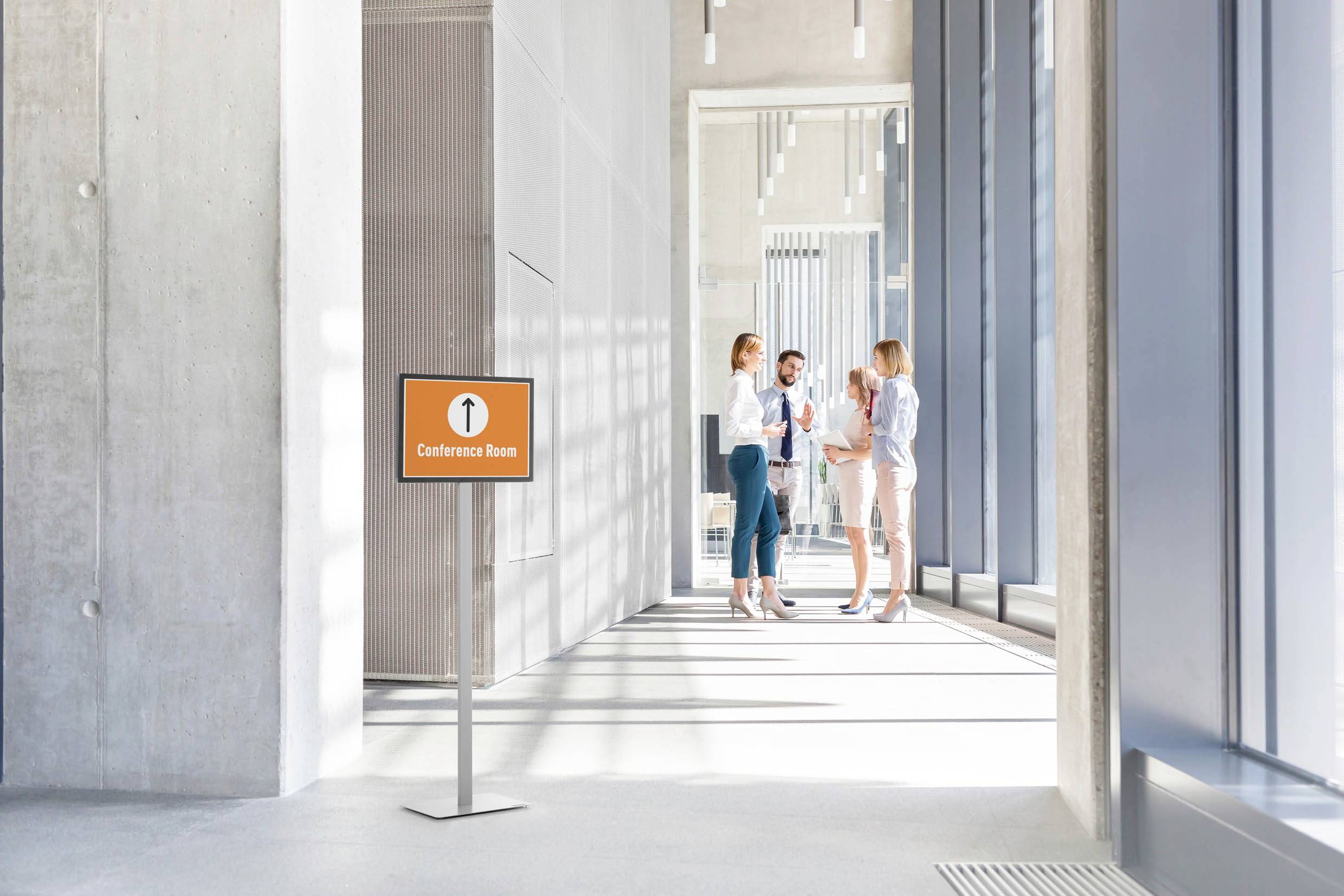 Drie personen praten met elkaar in een moderne kantoorgang. Een bord aan de linkerkant wijst de richting naar de 'vergaderzaal'.