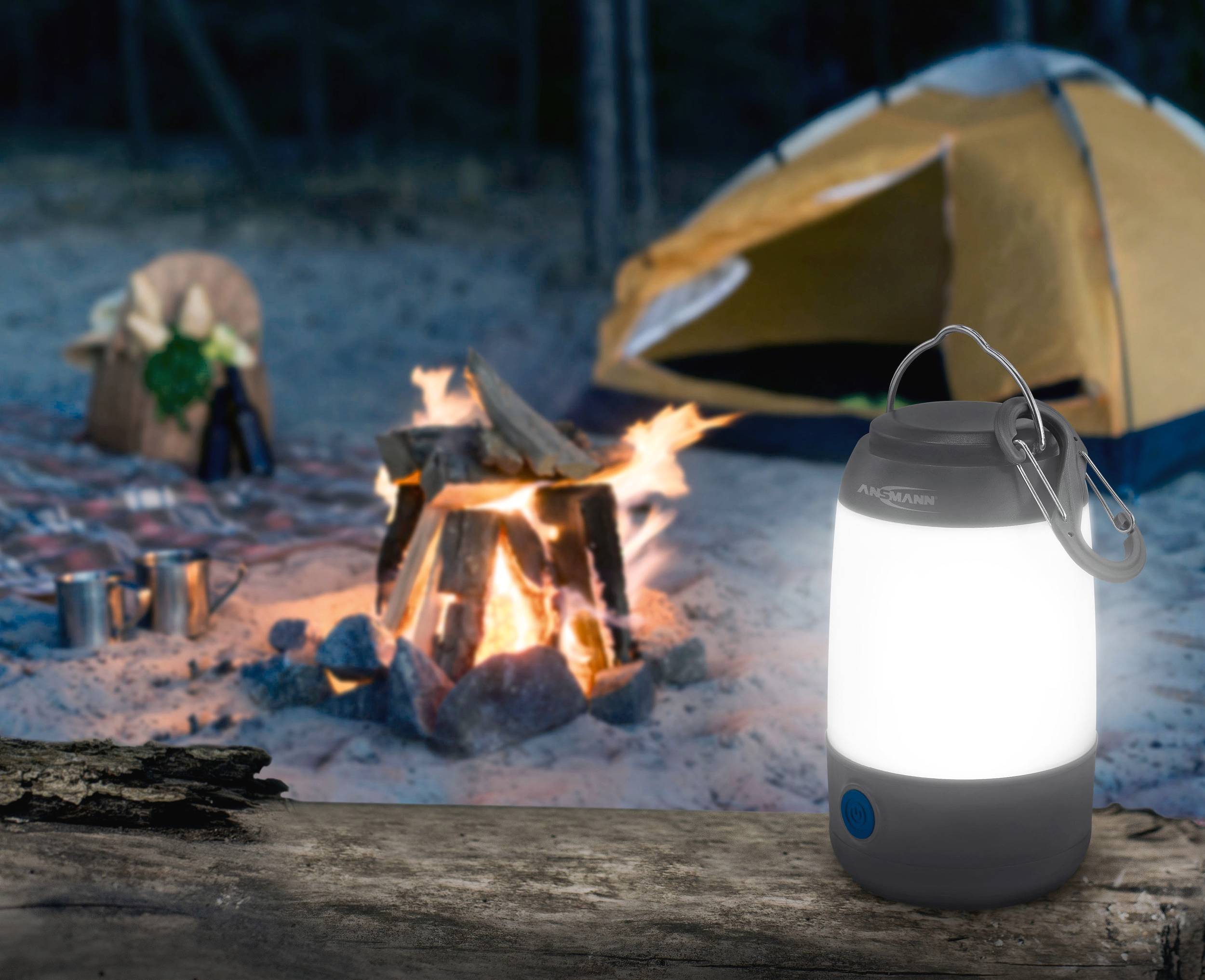 Kampeerterrein-tafereel met een verlicht kampvuur, tent op de achtergrond en een heldere lantaarn op een boomstam op de voorgrond.