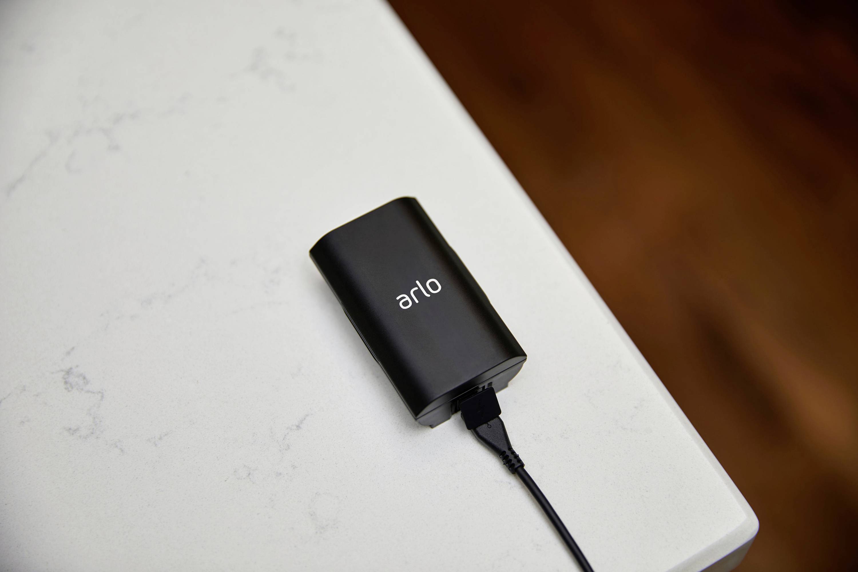Zwarte accu met 'arlo'-opschrift ligt op een witte tafel, aangesloten op een oplaadkabel.