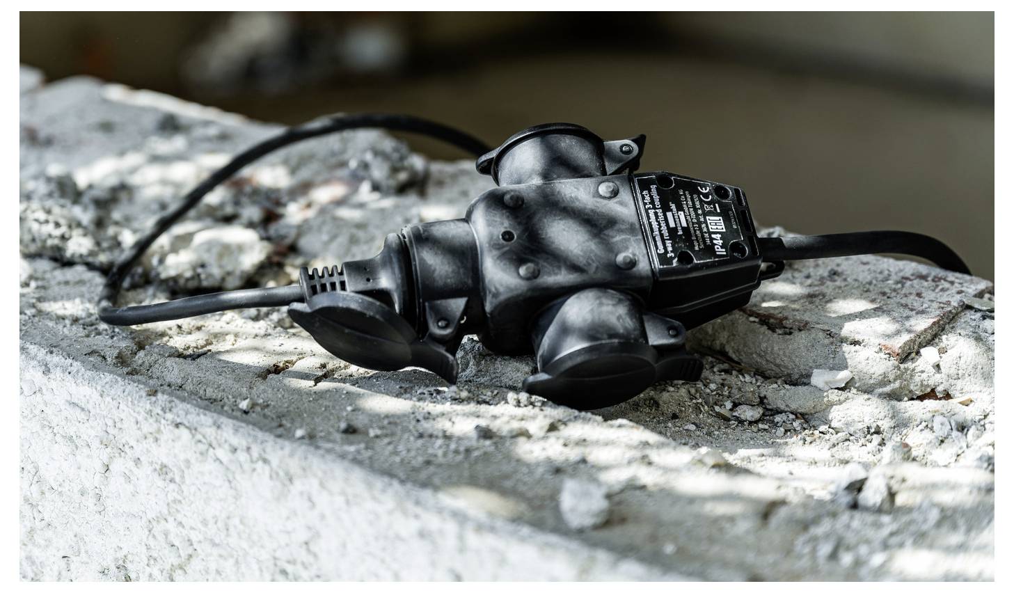 Zwarte elektrische stekkeradapter liggend op een ruw betonnen oppervlak buiten, met een aansluitkabel eraan bevestigd en een etiket zichtbaar bovenaan.