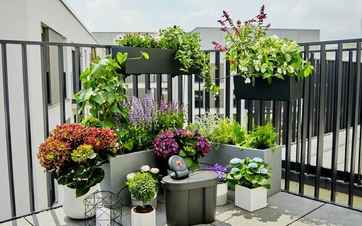Verschillende kleurrijke planten en bloemen in potten op een balkon met een metalen balustrade en een modern gebouw op de achtergrond.