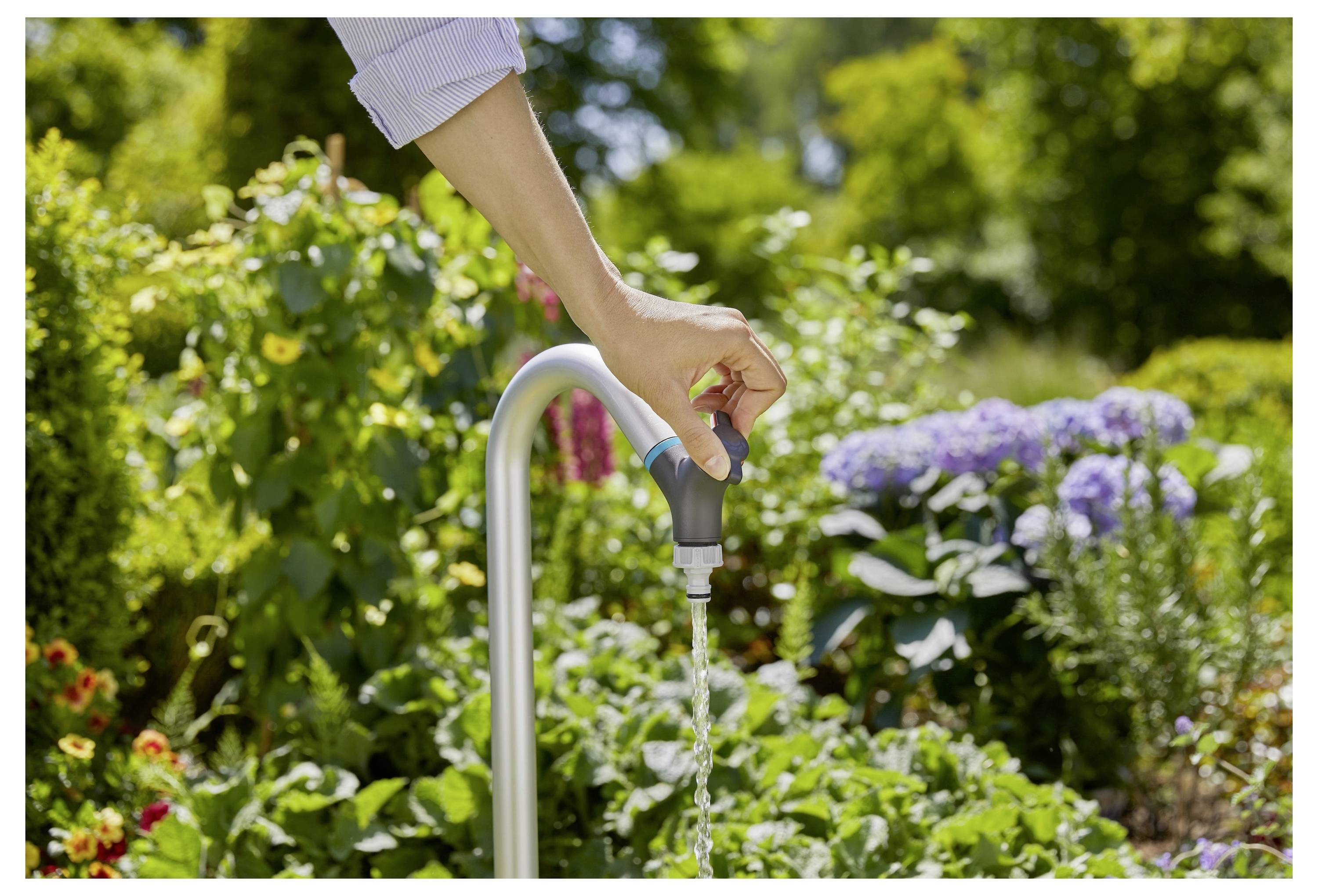 Een persoon zet een tuinkraan aan en besproeit planten die omringd zijn door bloeiende bloemen in een levendige tuin.