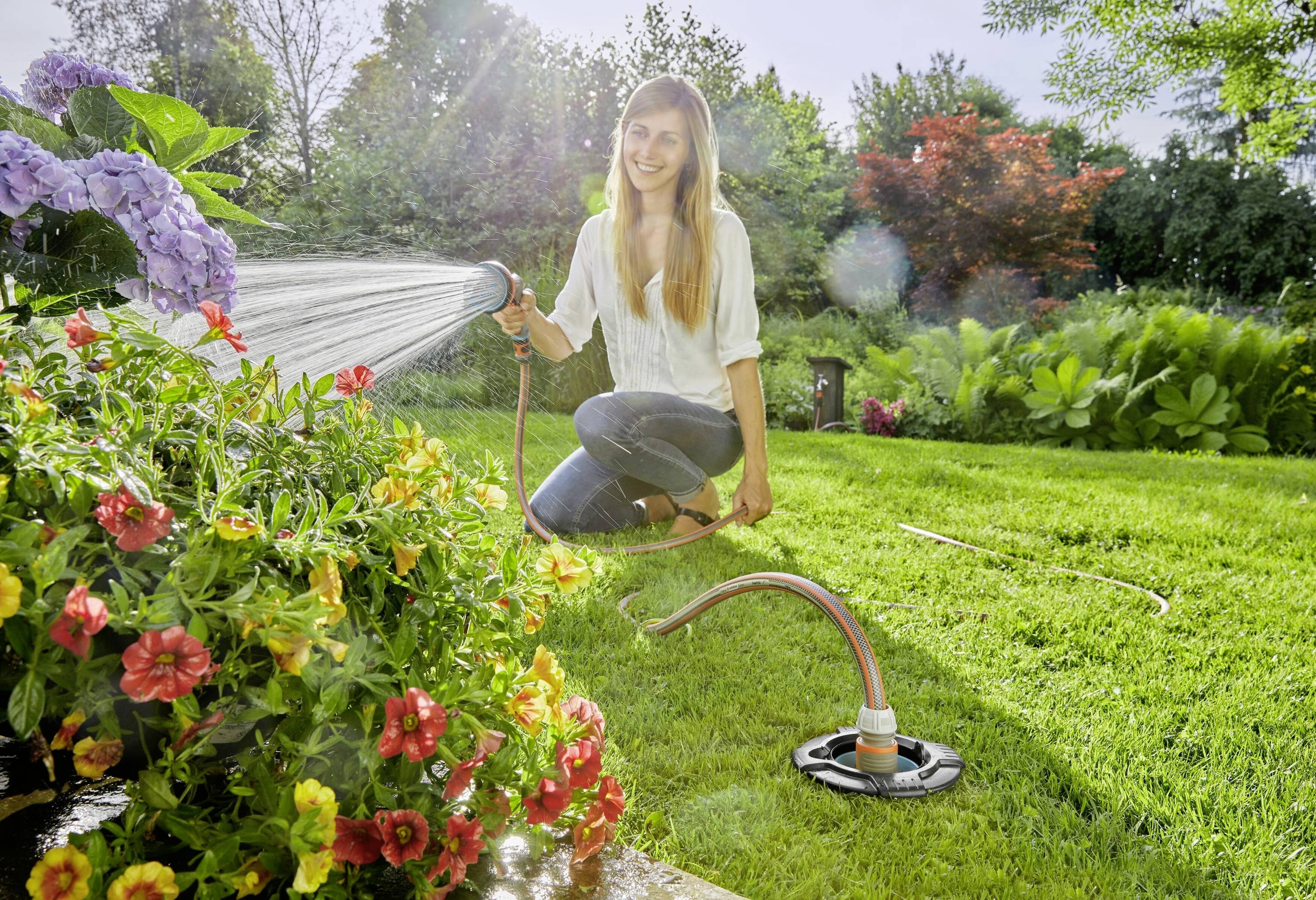 Een lachende vrouw water geeft kleurrijke bloemen met een tuinslang in een zonnige tuin. Op de achtergrond zijn bomen zichtbaar.