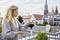 Een vrouw staat op een terras en houdt een voorwerp in haar hand. Op de achtergrond is een stadslandschap met een kerk zichtbaar.