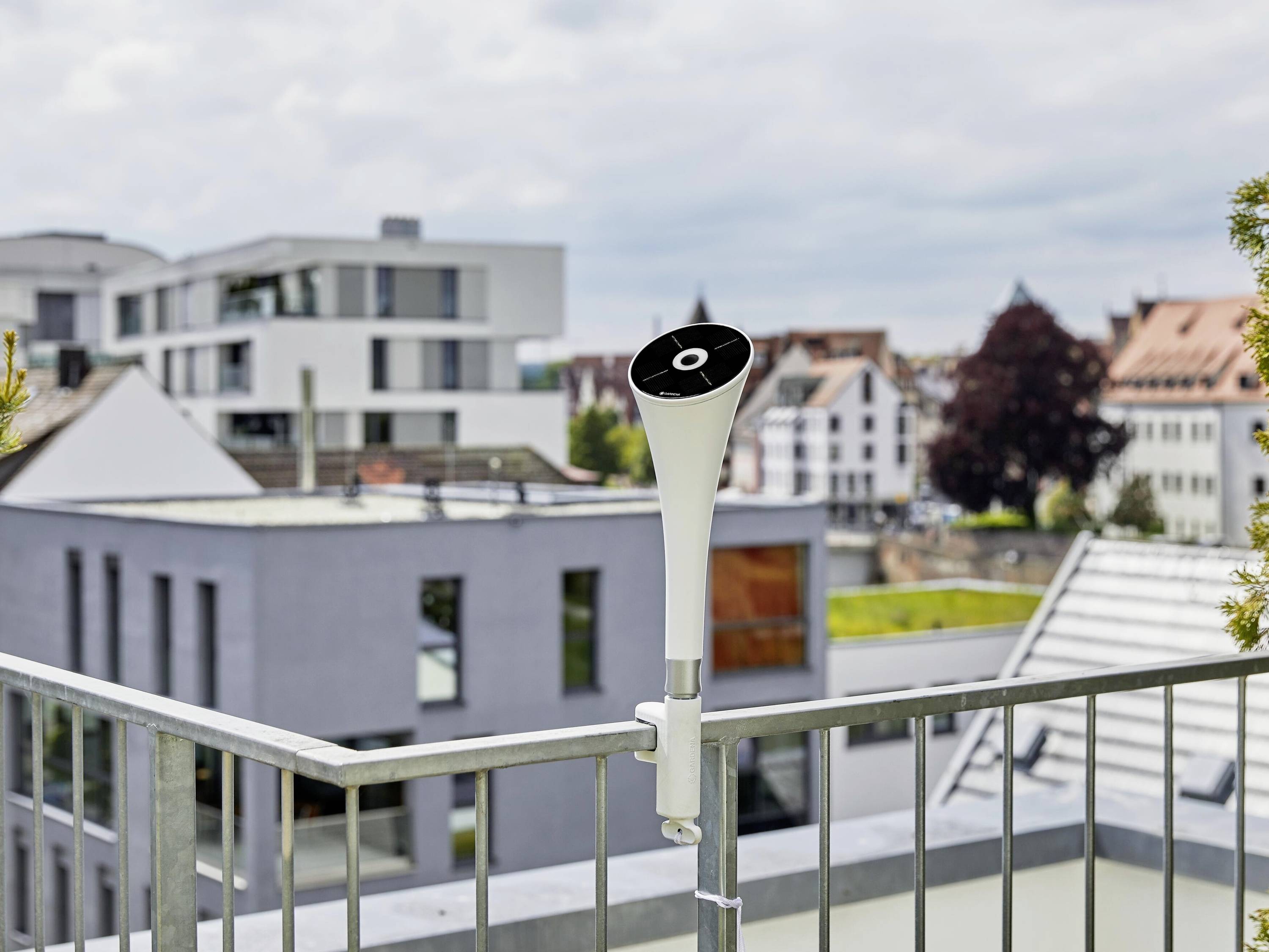 Een witte weersstation is geïnstalleerd op een balkon, omgeven door moderne gebouwen in een stedelijke omgeving.