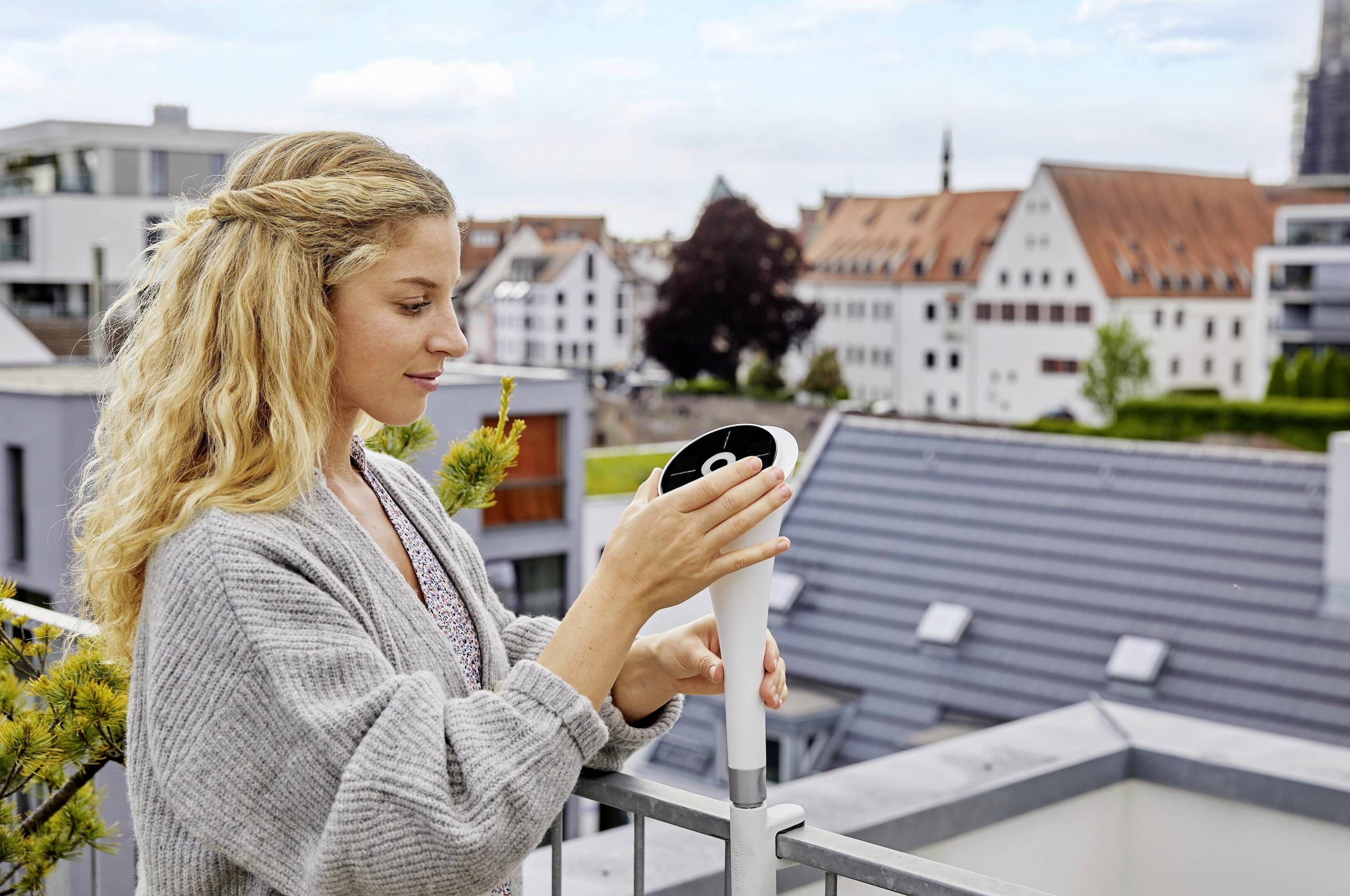 Een vrouw monteert een buitencamera op een balkon. Op de achtergrond zijn moderne gebouwen en een bewolkte lucht te zien.
