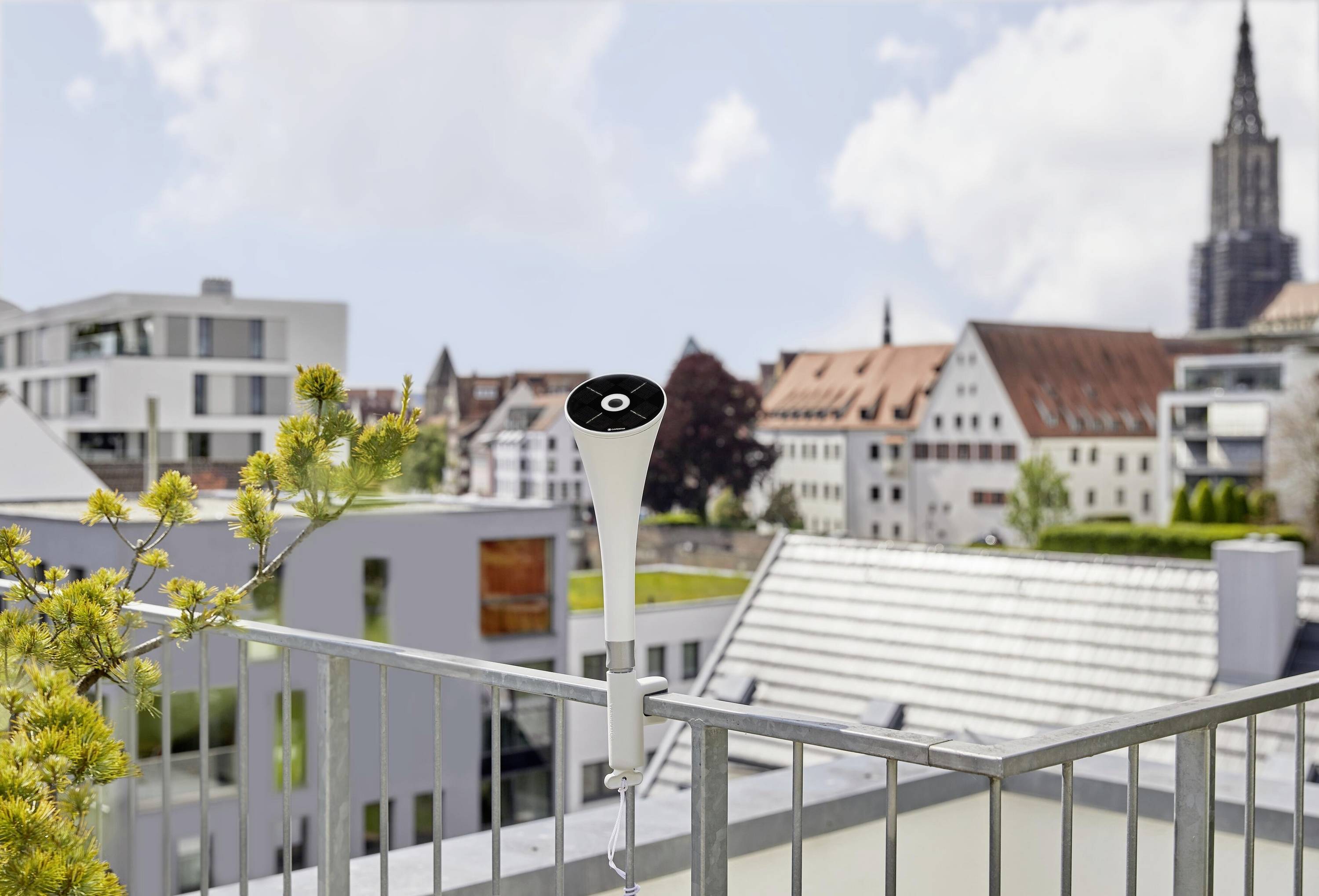 Een witte, weersbestendige luidspreker op een balkon met uitzicht op een stedelijk landschap, met moderne en historische gebouwen eronder.