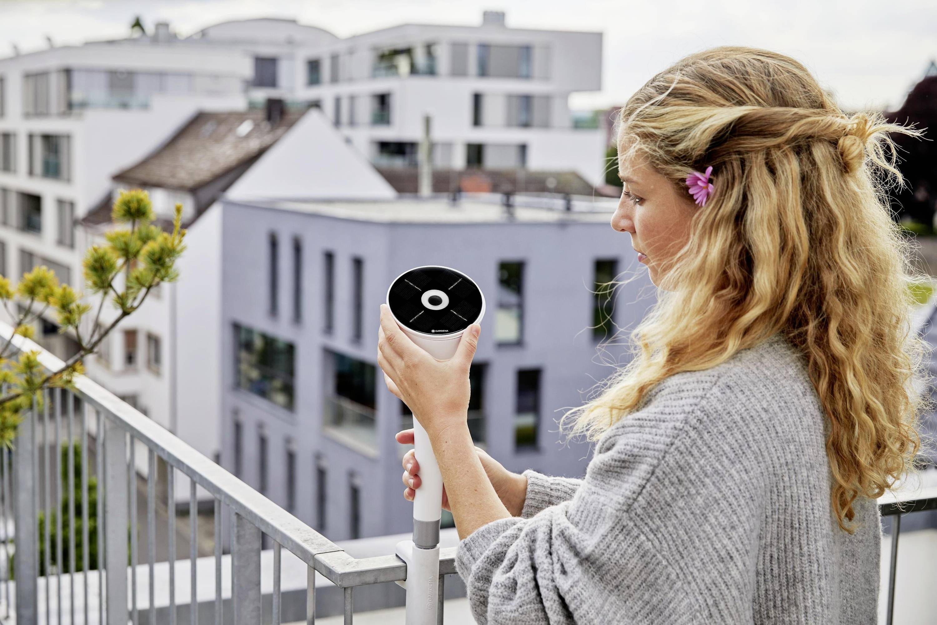 Een vrouw houdt een modern bewakingsapparaat op een balkon, met stedelijke architectuur op de achtergrond, in een rustige woonomgeving.