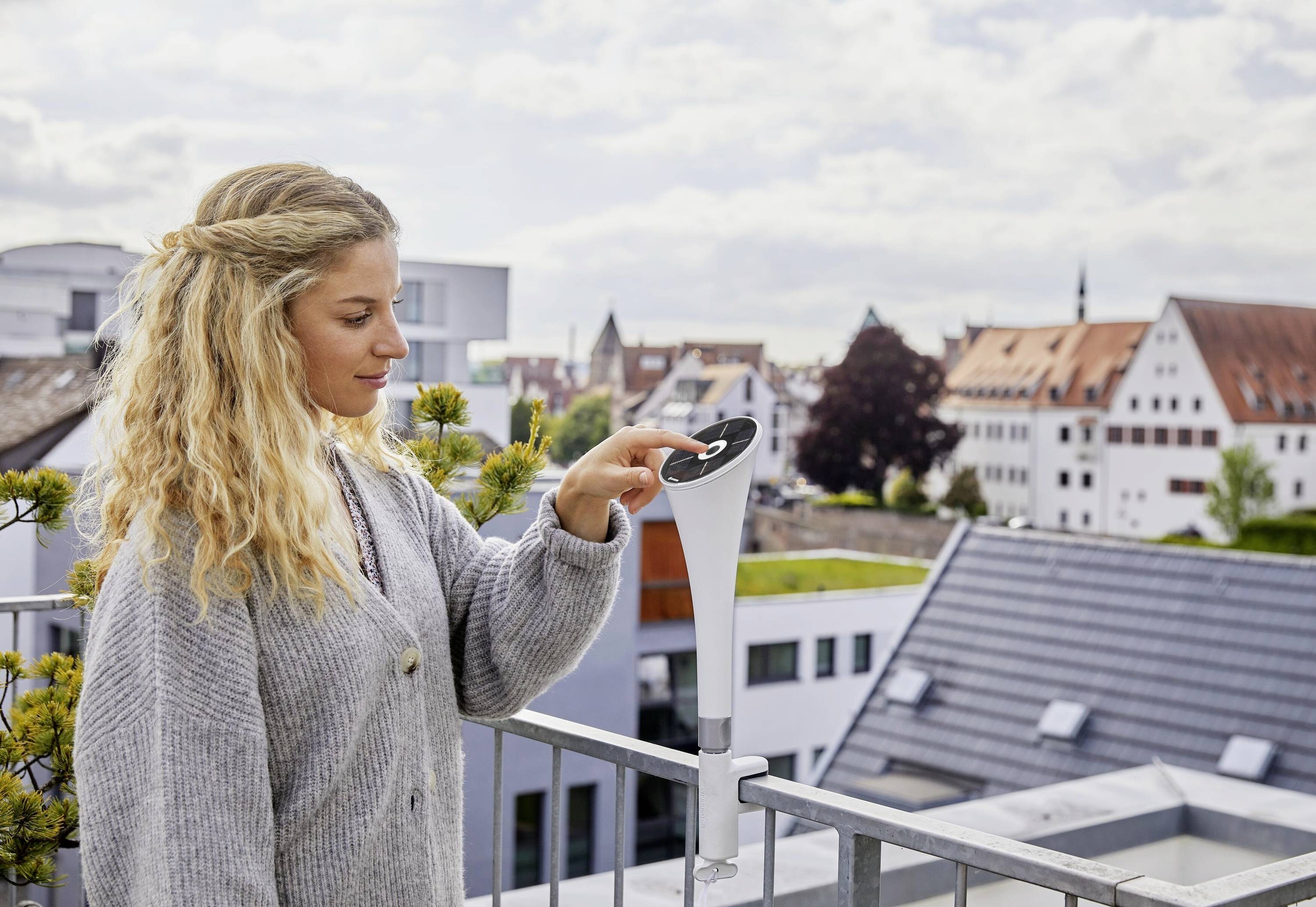 Een persoon op een balkon bedient een apparaat, mogelijk een weermeetinstrument, met een stad op de achtergrond.