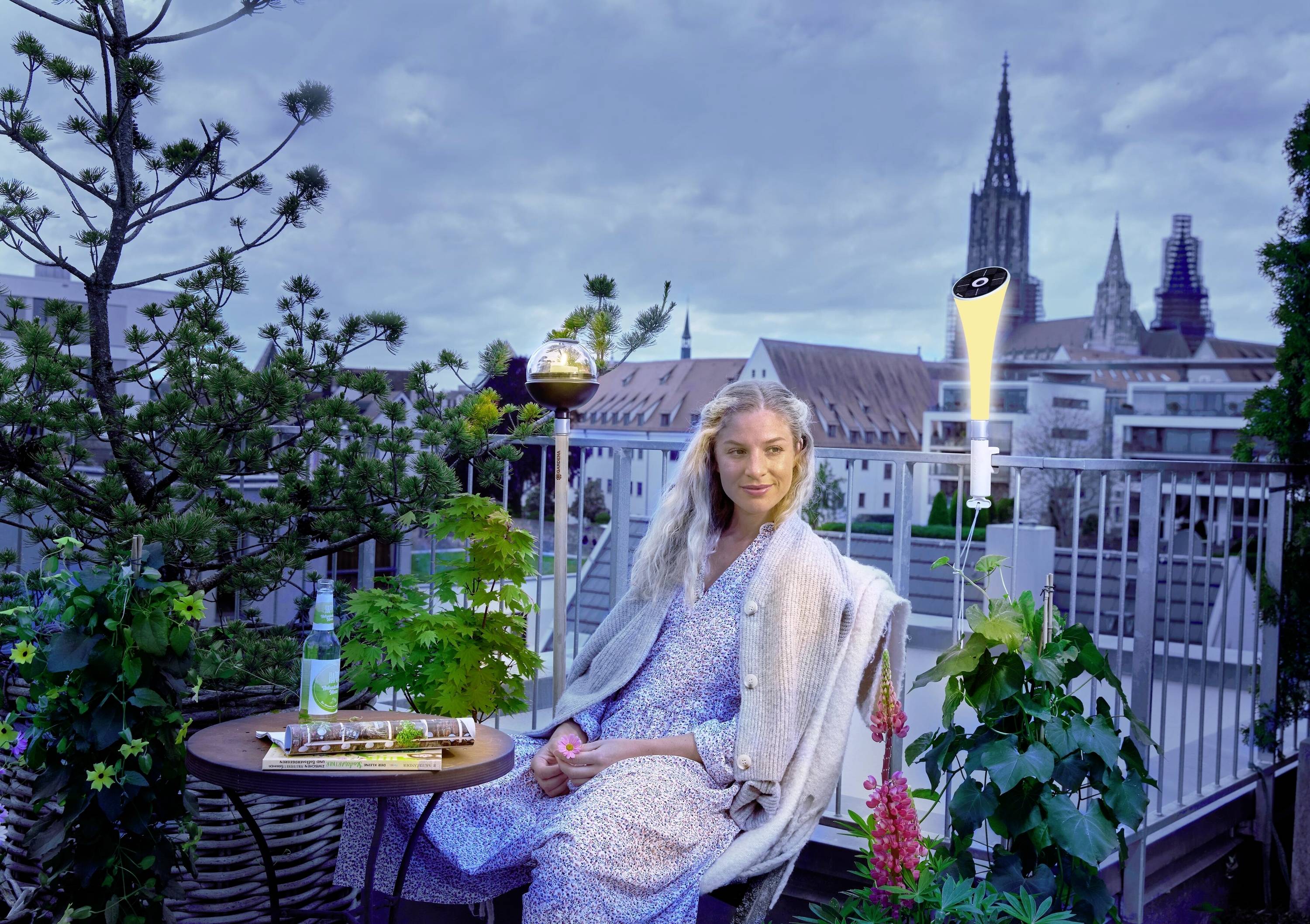 Een vrouw zit ontspannen op een balkon, omringd door planten bij het invallen van de avond. Op de achtergrond zijn gebouwen en een kerktoren zichtbaar.