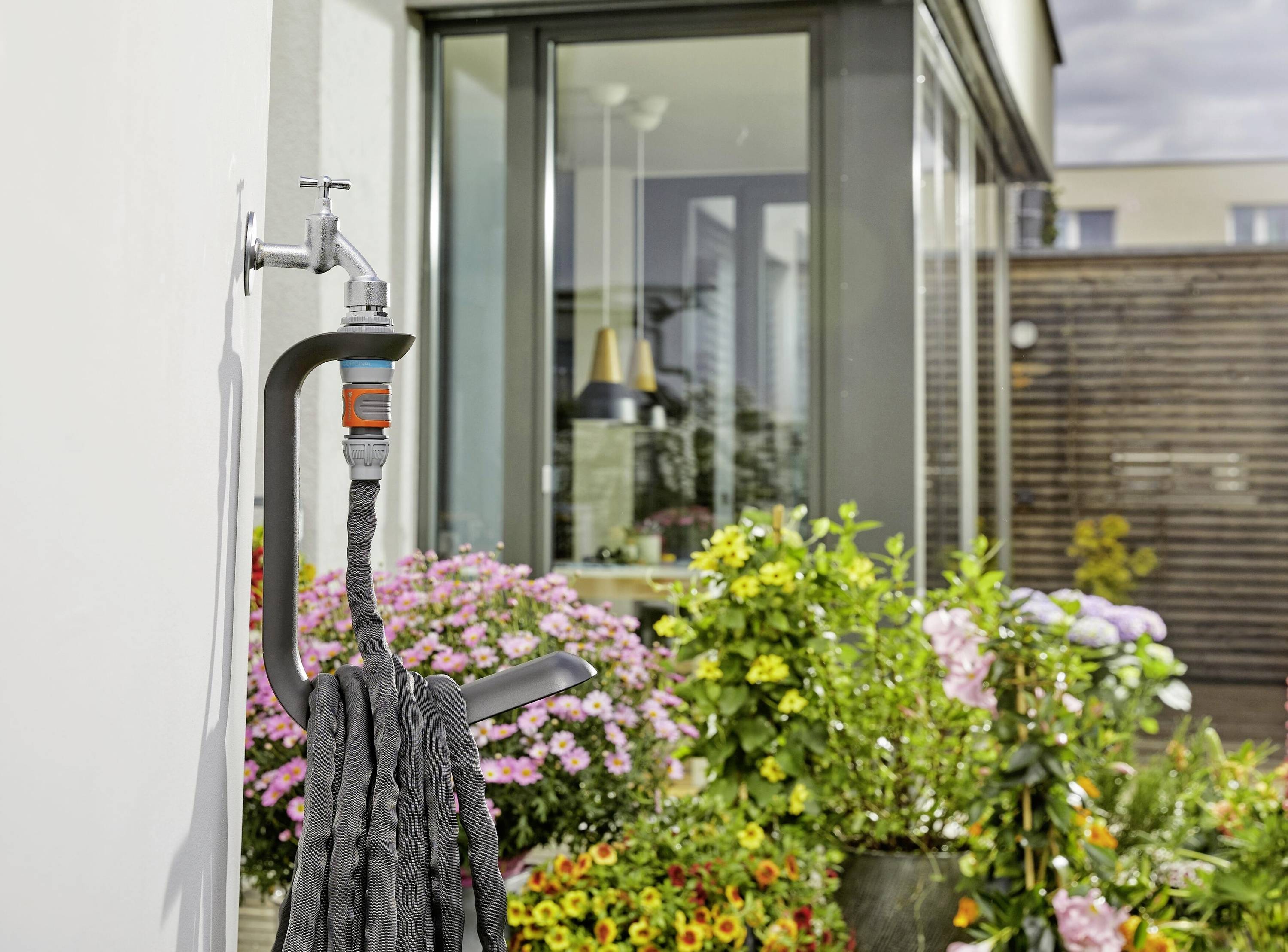 Een waterkraan met een slang eraan bevestigd hangt aan de muur. Op de achtergrond bloeien bloemen en staat een modern gebouw.