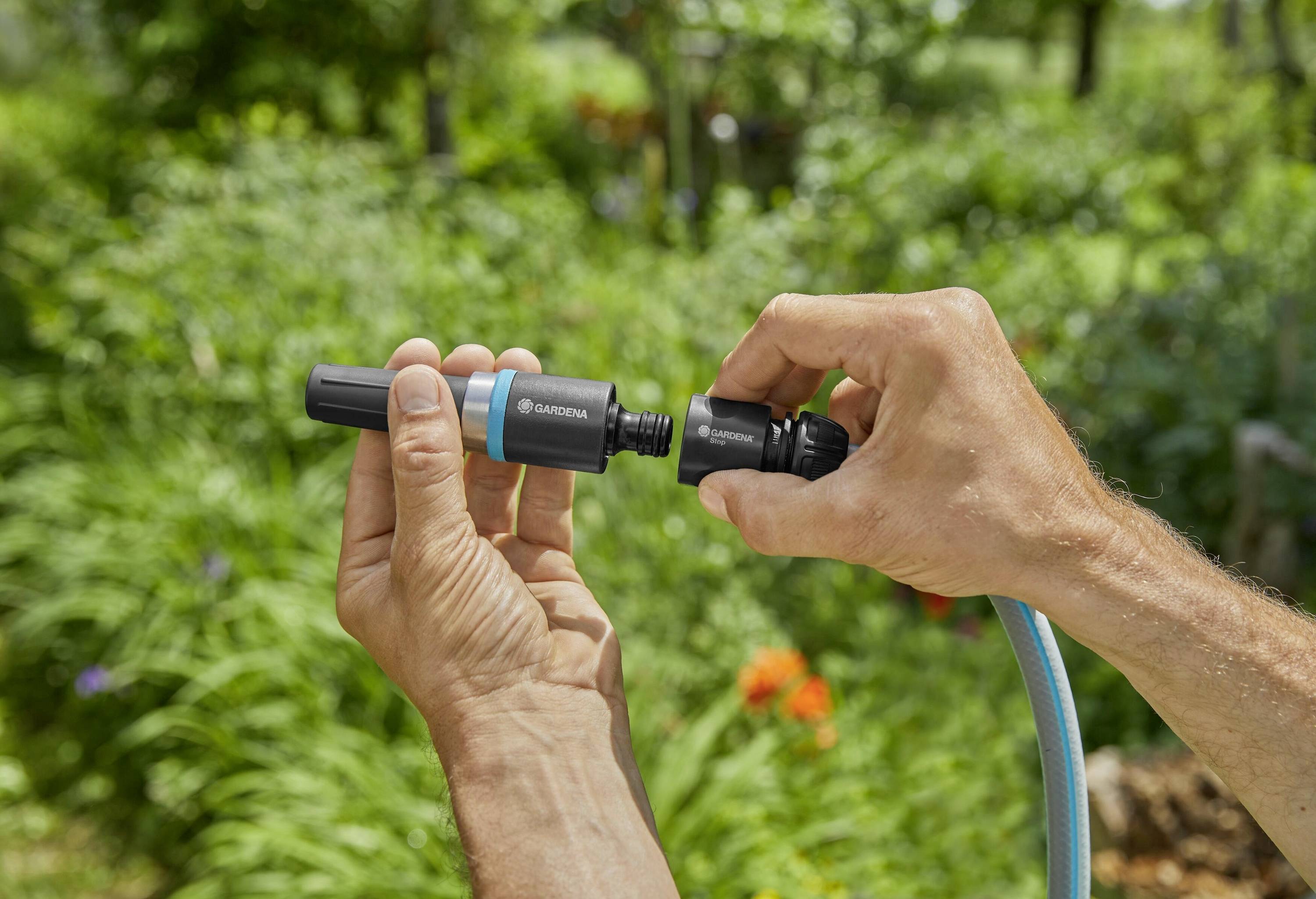 Twee handen houden en verbinden onderdelen van een tuinslangaansluiting, met een onscherpe groene tuinachtergrond buiten.