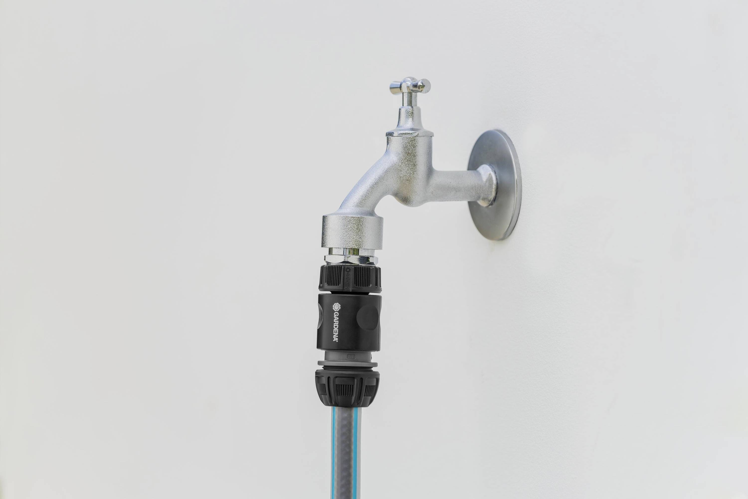 Een waterkraan aan een muur met een aangesloten zwarte adapter en blauwe slang, klaar voor watervoorziening buiten.