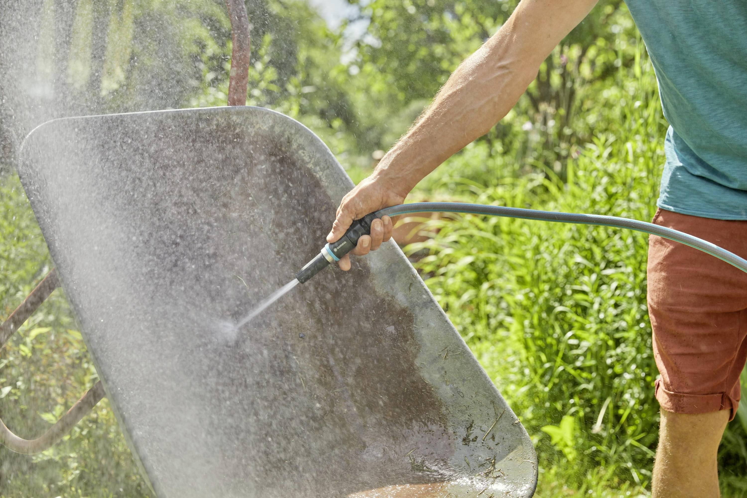 Een persoon reinigt een kruiwagen met een waterslang in de tuin. Op de achtergrond zijn groene planten zichtbaar.