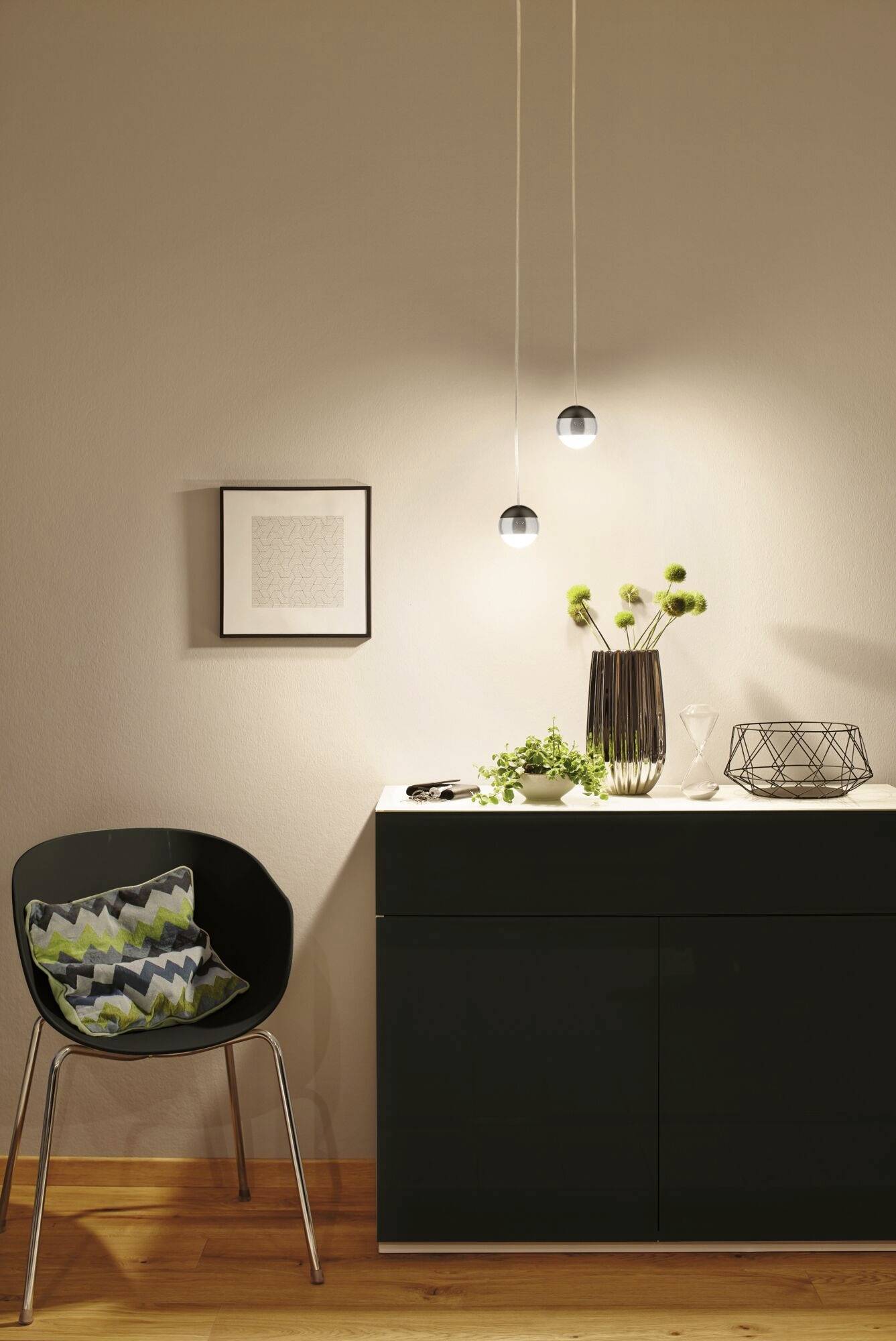 Modern interieur met zwarte stoel, decoratief kussen, kast, planten en hangende lampen in een elegant verlichte ruimte.