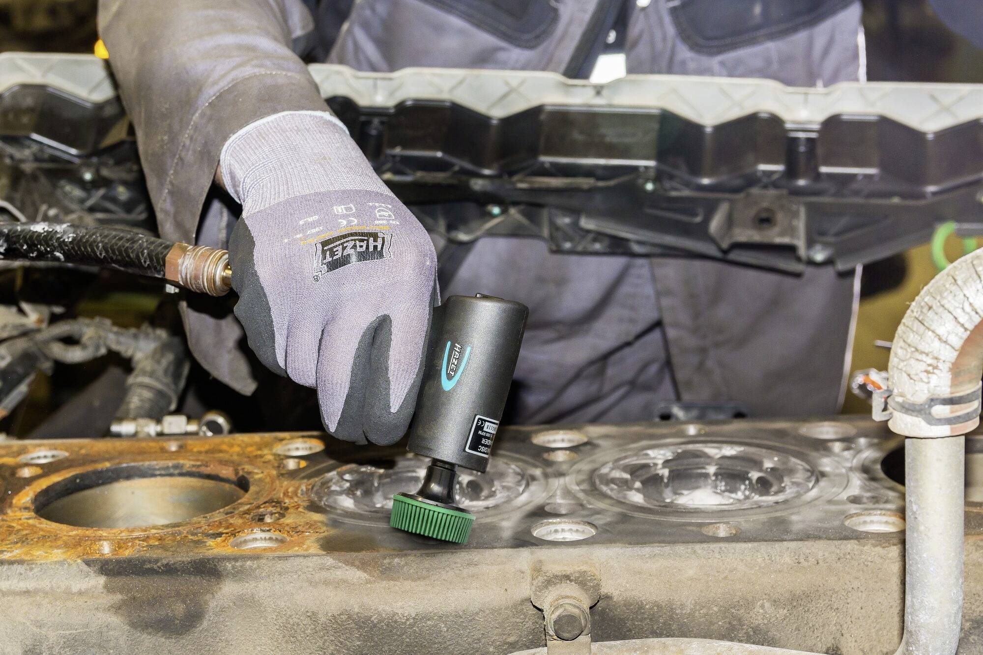 Een monteur reinigt het motorblok van een voertuig met een elektrisch slijpgereedschap in een werkplaats.