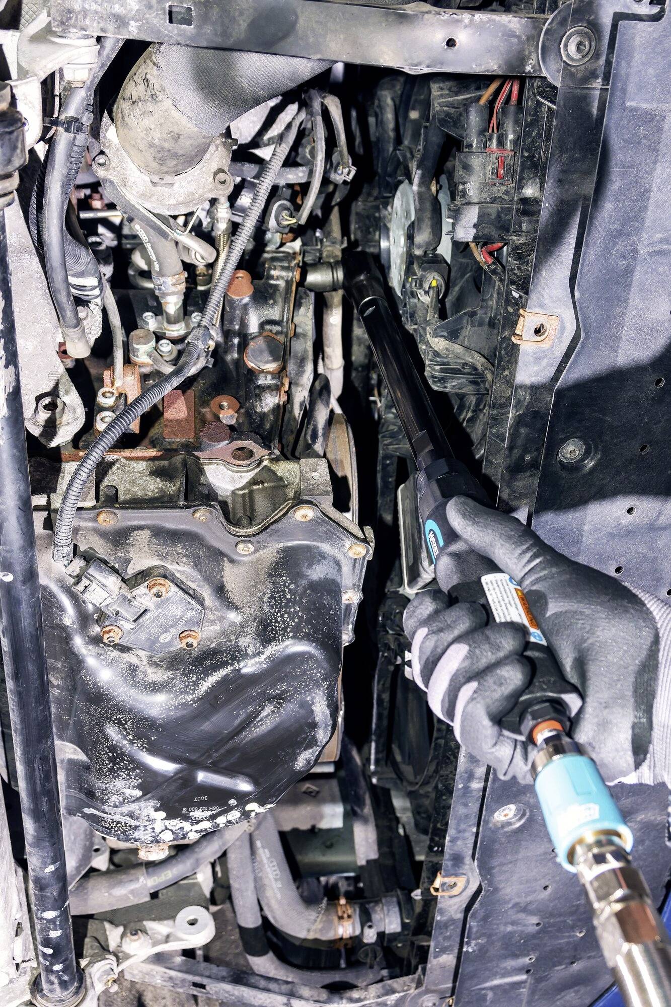 Close-up van een monteur met handschoenen die met een gereedschap werkt aan een open automotor, omringd door kabels en onderdelen.