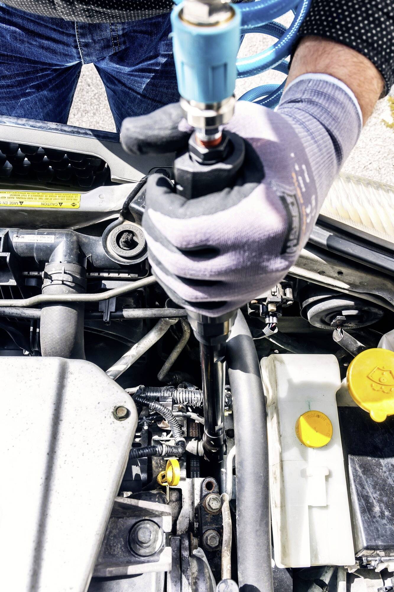Een persoon repareert een automotor met een pneumatisch gereedschap. Ze draagt werkhandschoenen. De motorruimte is gedeeltelijk zichtbaar.
