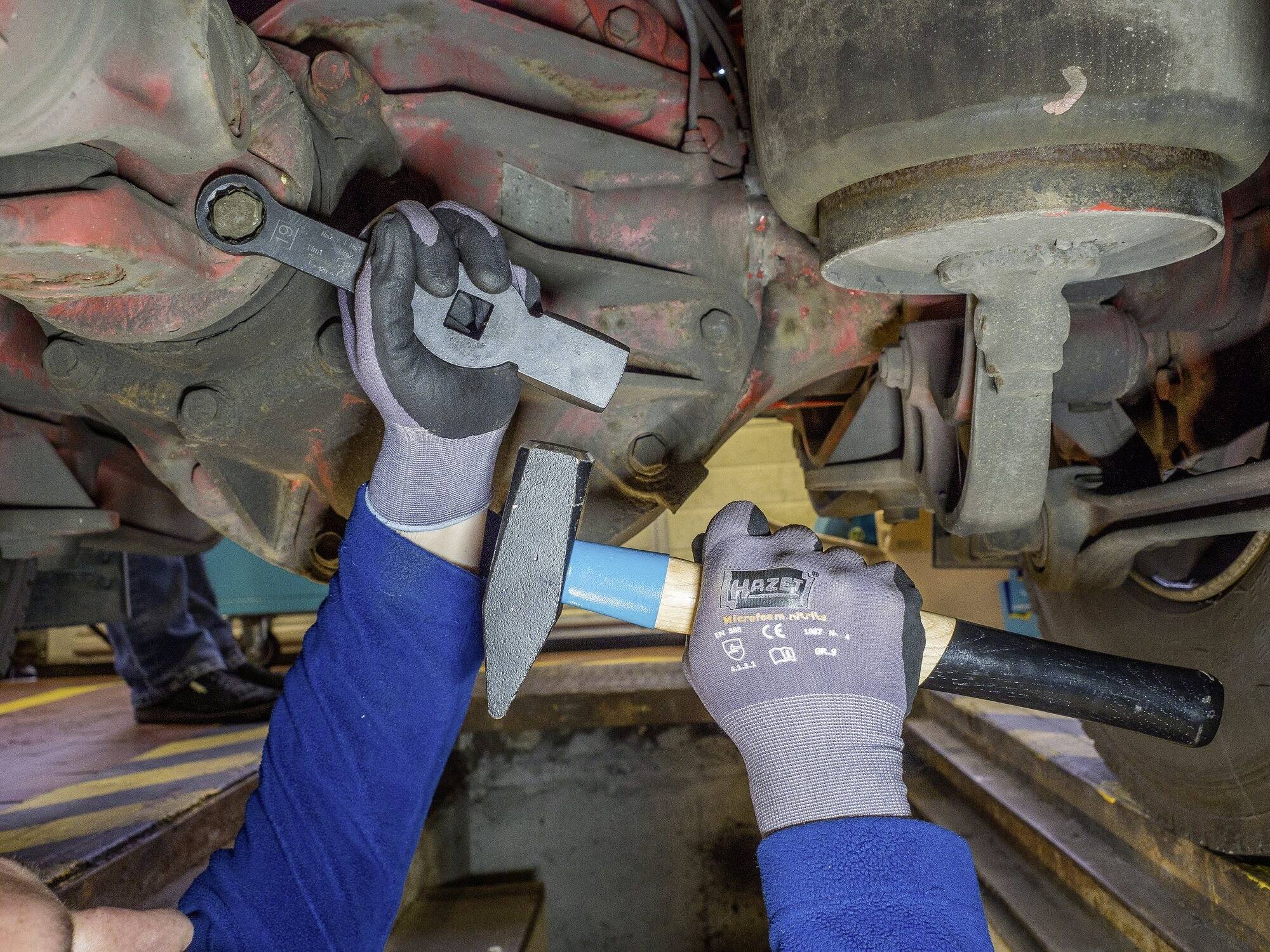 Monteur repareert onderkant van een voertuig in een werkplaats, houdt moersleutel en hamer vast.