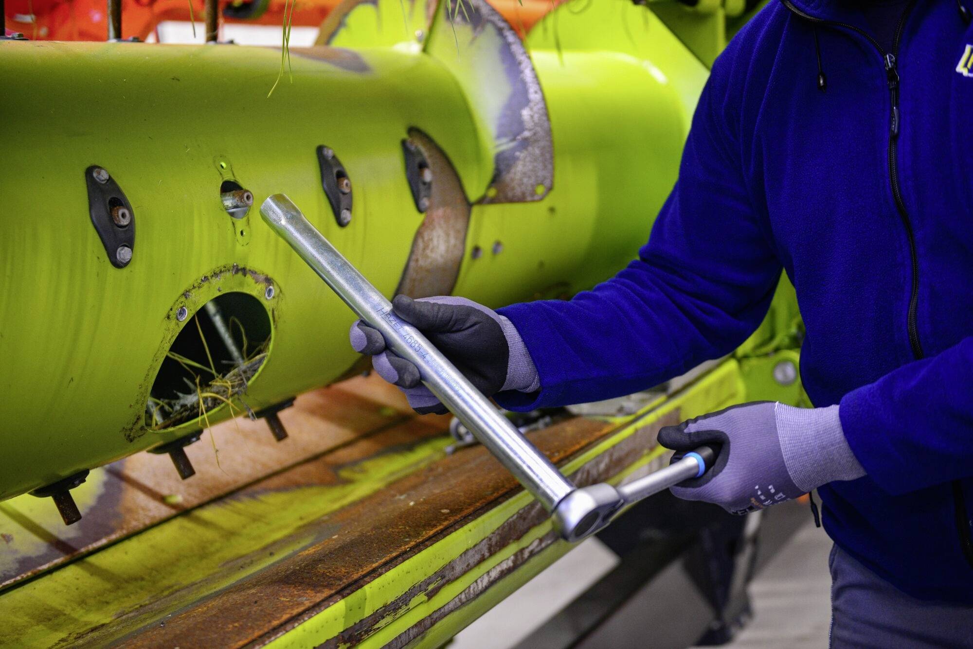 Een persoon in blauwe kleding repareert een grote groene machine met een gereedschap. De omgeving lijkt industrieel te zijn.
