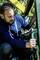 Man met baard in blauwe hoodie repareert een groen metalen leuningwerk met een gereedschap buiten.