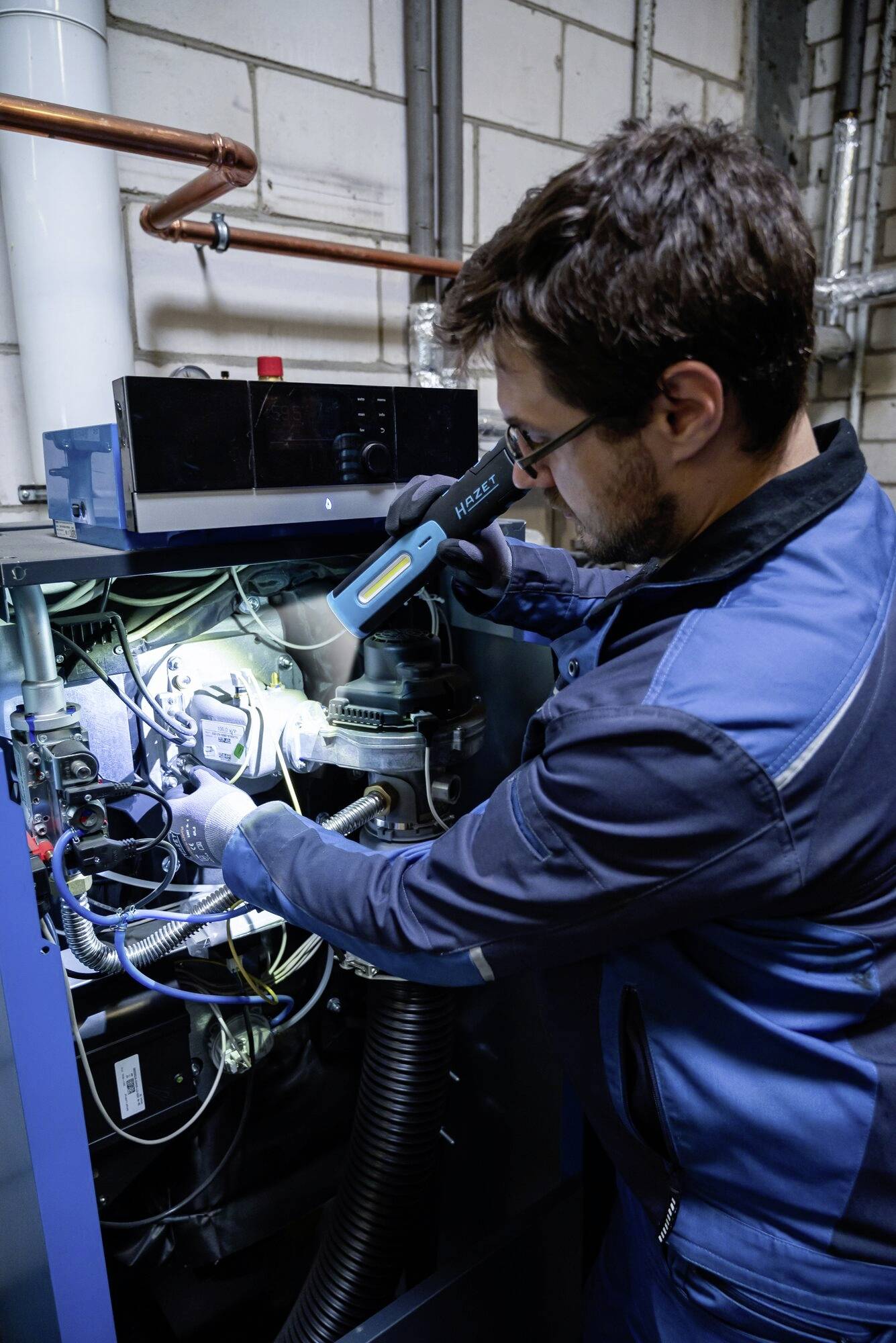 Een persoon in blauwe werkkleding repareert een complex verwarmingssysteem in een technische ruimte, die verbonden is met buizen en kabels.