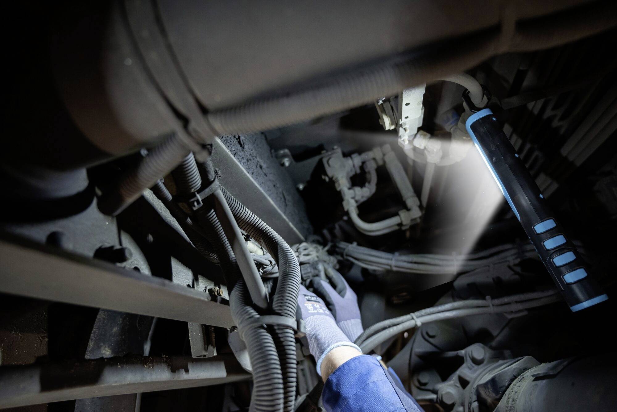 Een persoon werkt met handschoenen en zaklamp onder een machine. Er zijn veel kabels en buizen zichtbaar.