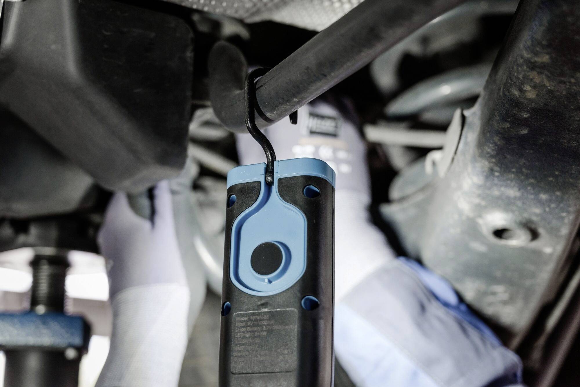 Monteur repareert voertuig, houdt een draagbare lamp onder de auto. Hij draagt handschoenen en gebruikt professioneel gereedschap.