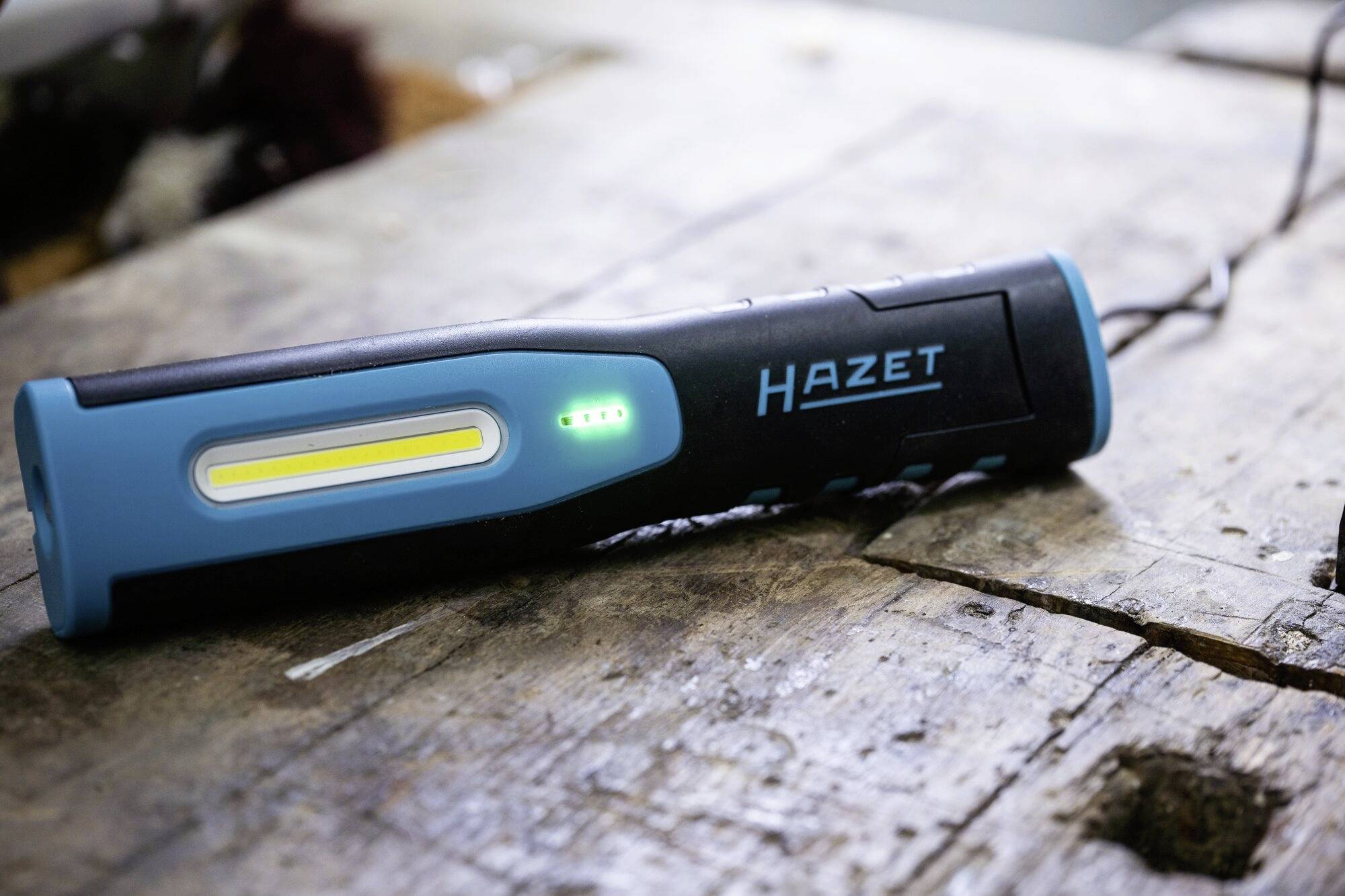 Een blauwe en zwarte werkverlichting van HAZET ligt op een houten tafel. De lamp is ingeschakeld en toont een groene laadstatus.