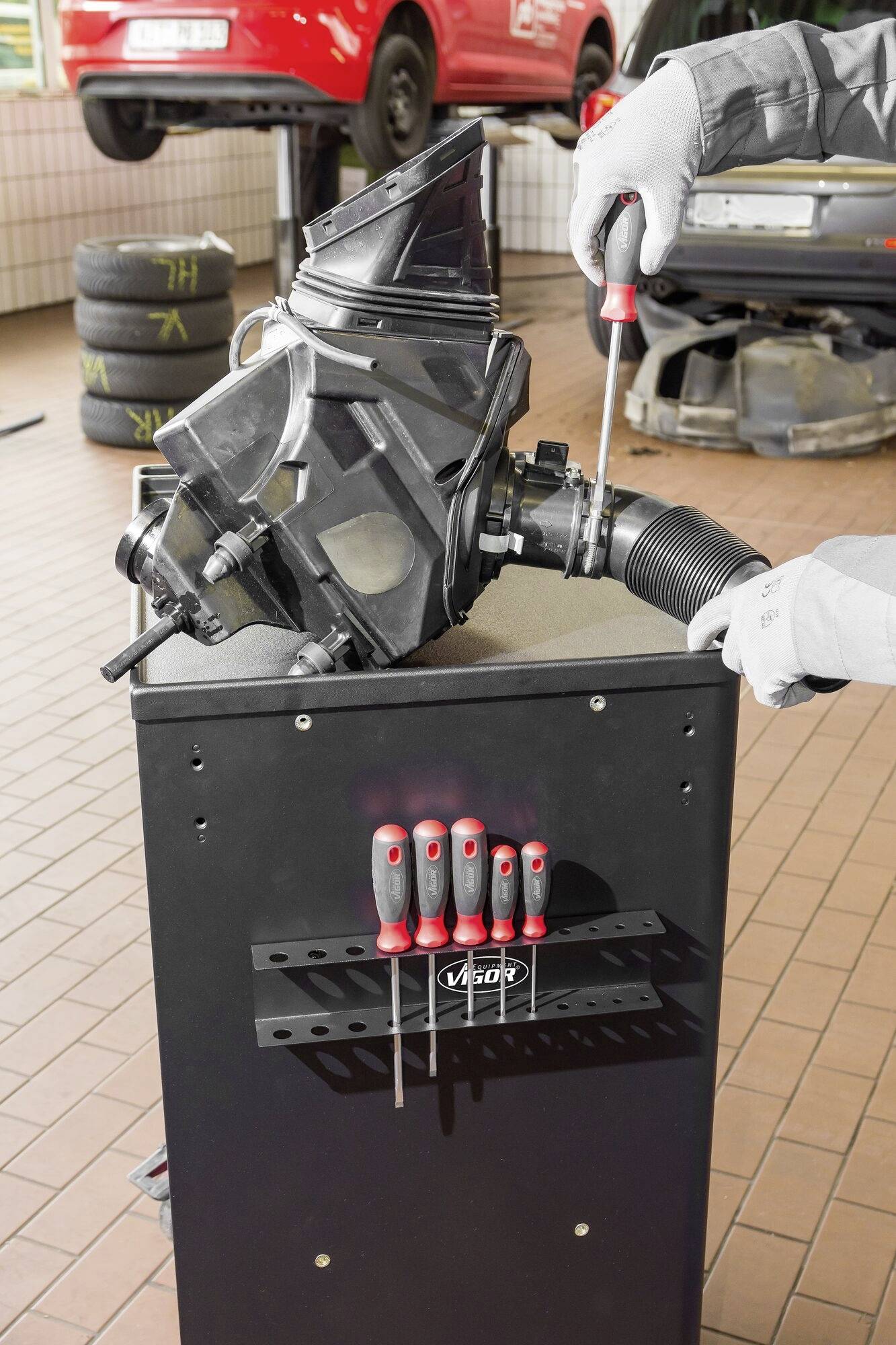 Een monteur werkt in een autogarage aan een onderdeel op een gereedschapswagen. Op de achtergrond zijn autobanden zichtbaar.