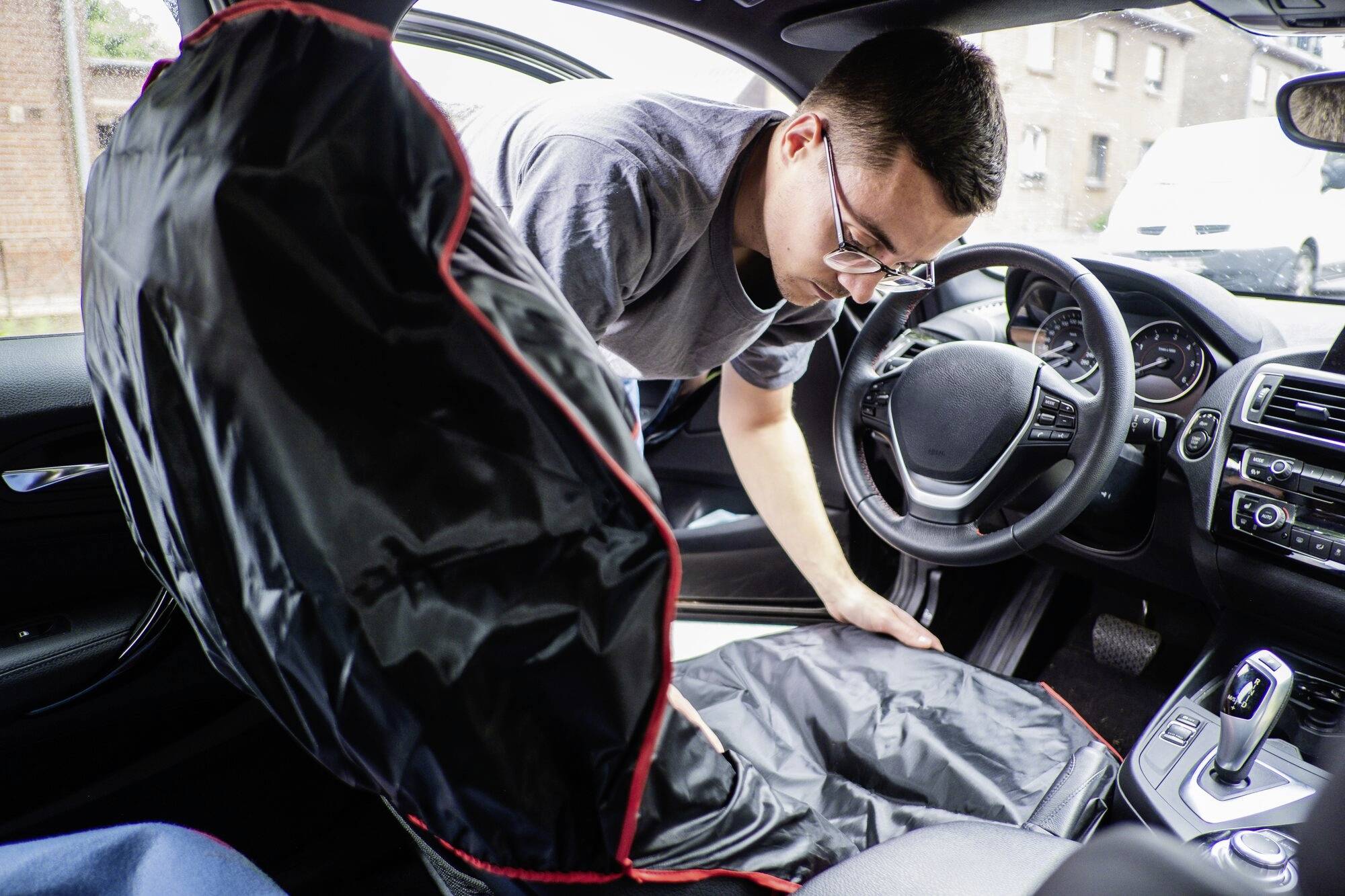 Een man in een auto legt een zwarte stoelhoes met rode rand op de bestuurdersstoel.