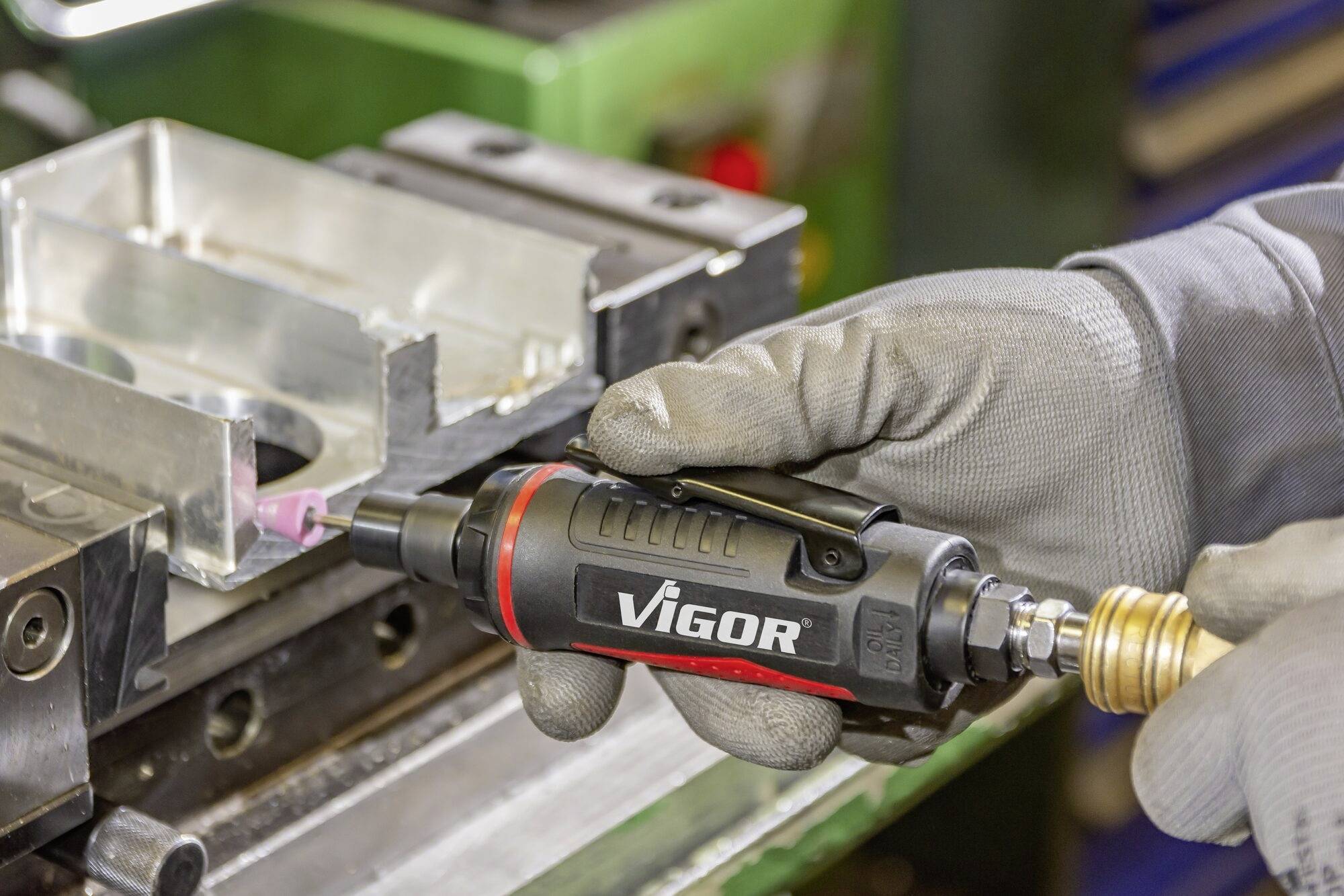 Een gehandschoende hand houdt een Vigor-gereedschap vast en bewerkt een metalen onderdeel in een werkplaatsomgeving.