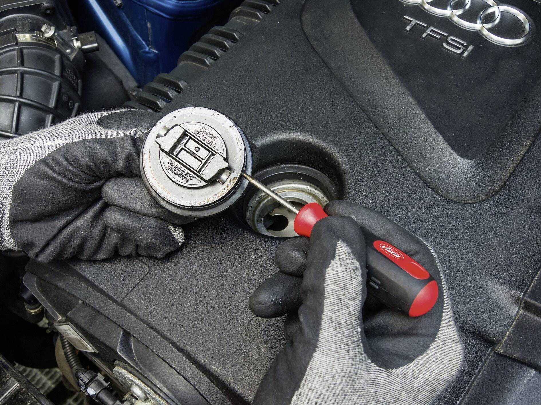 Close-up van handen met handschoenen die met een schroevendraaier de olievuldop van een motor openen. Motor met 'TFSI'-label.