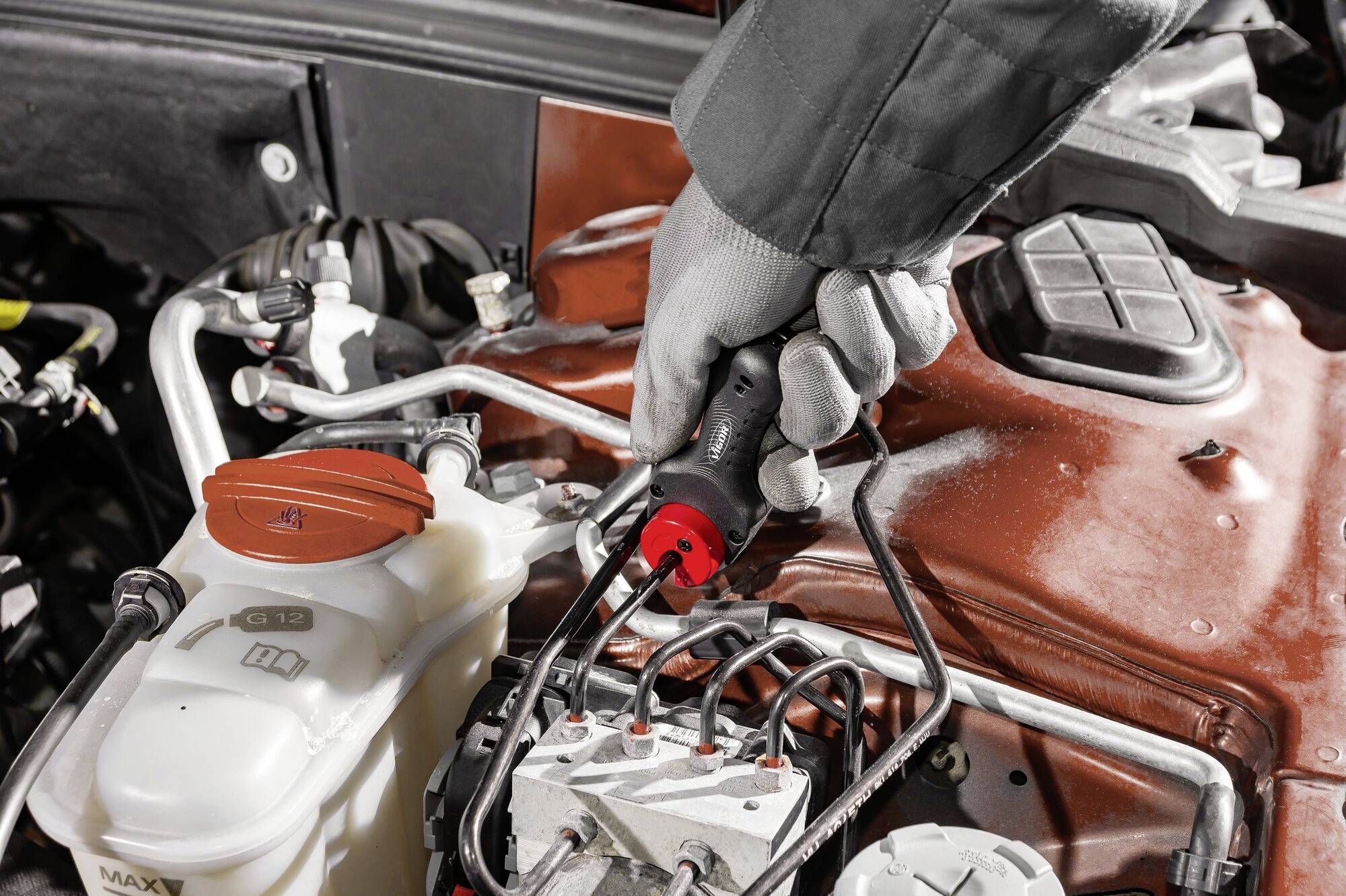 Een persoon met een grijze handschoen gebruikt een gereedschap om de motor van een auto te repareren. Op de voorgrond staat een container met vloeistof.
