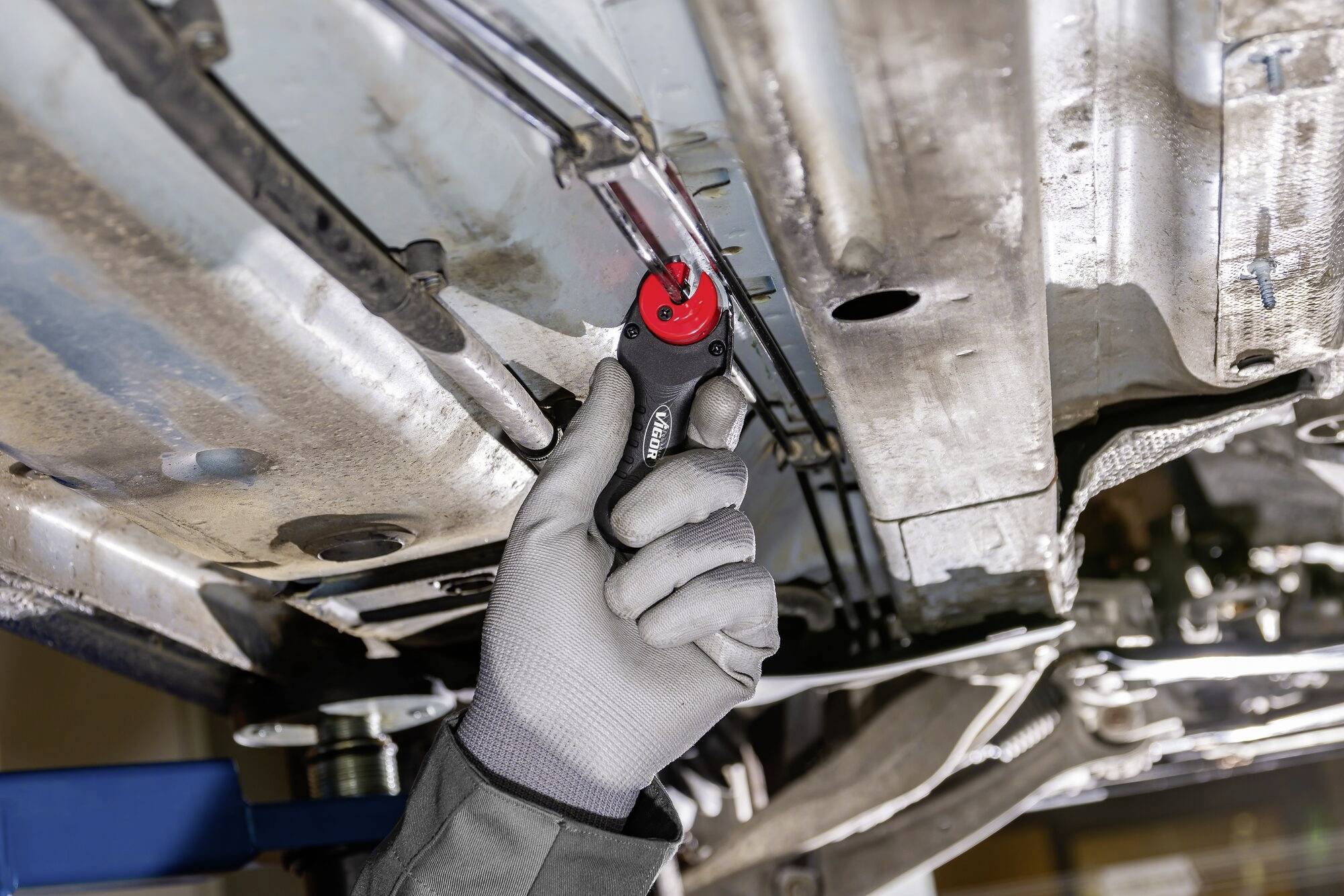 Een monteur met grijze handschoenen gebruikt een rood gereedschap om aan de onderkant van een voertuig te werken.