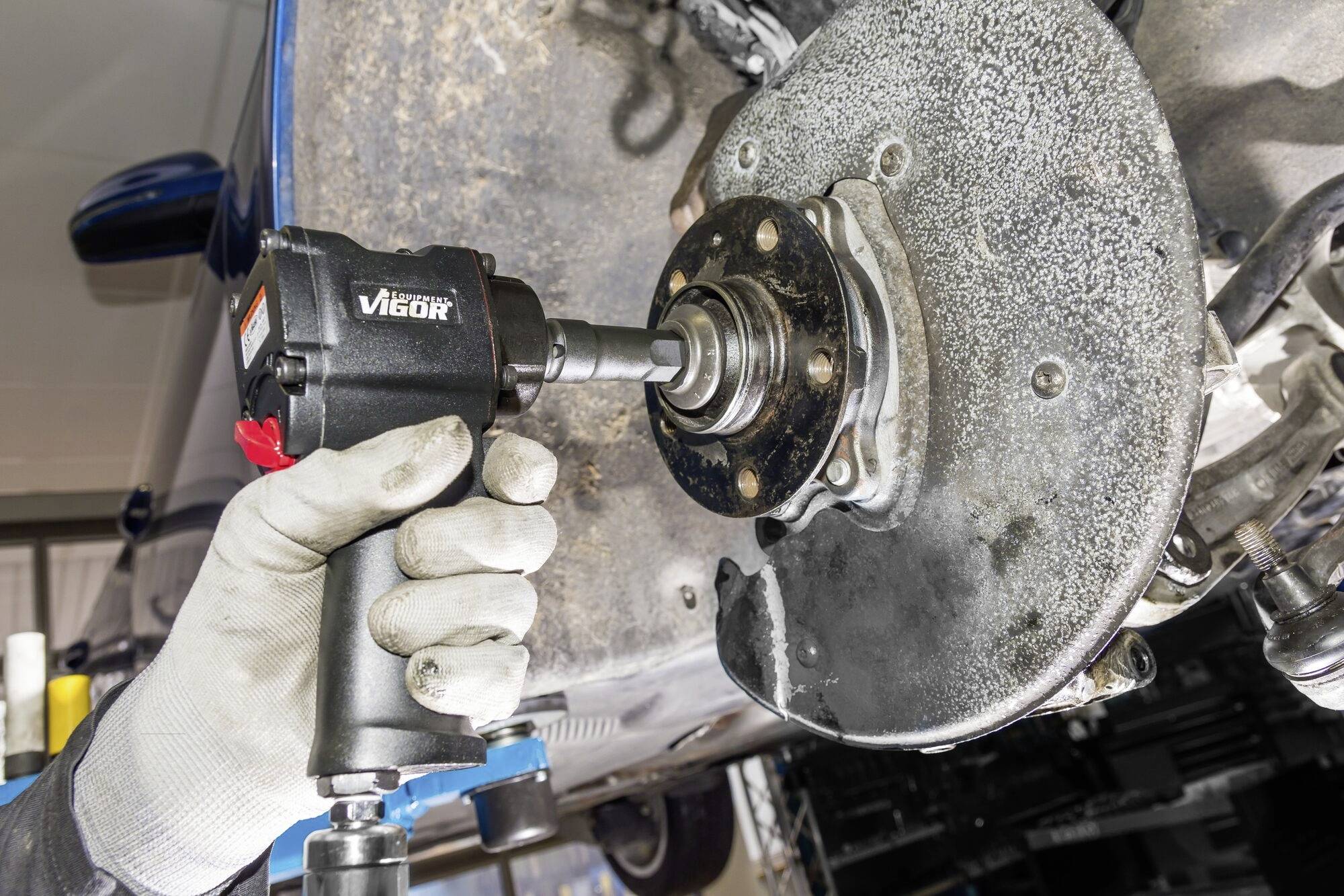 Een persoon met handschoenen repareert een auto en draait bouten vast met een elektrisch gereedschap aan een remschijf.