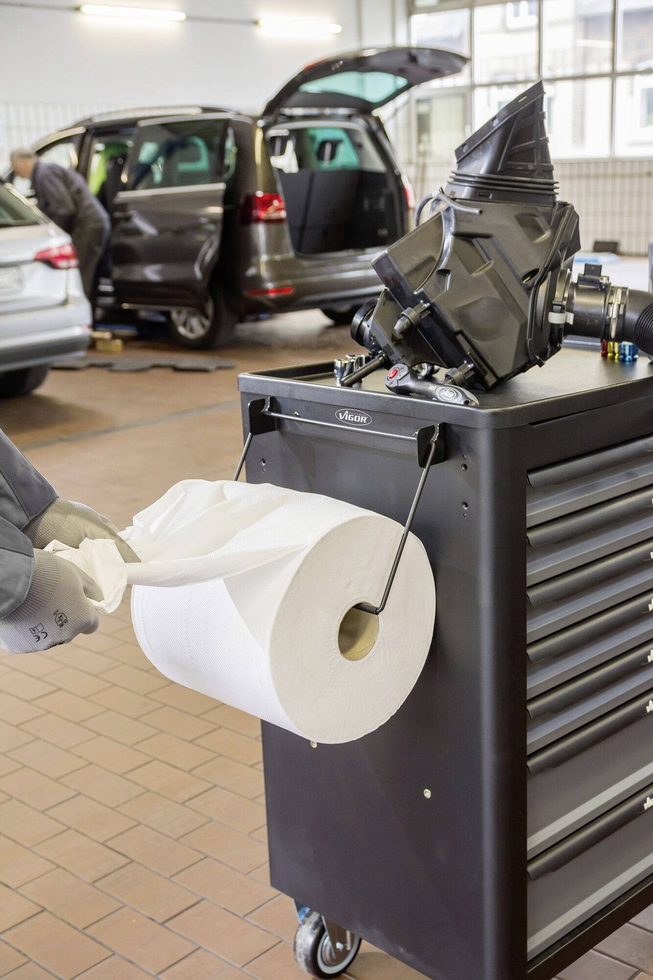 Een automonteur gebruikt papieren doeken bij een gereedschapswagen in een autogarage. Op de achtergrond wordt gewerkt aan een auto.